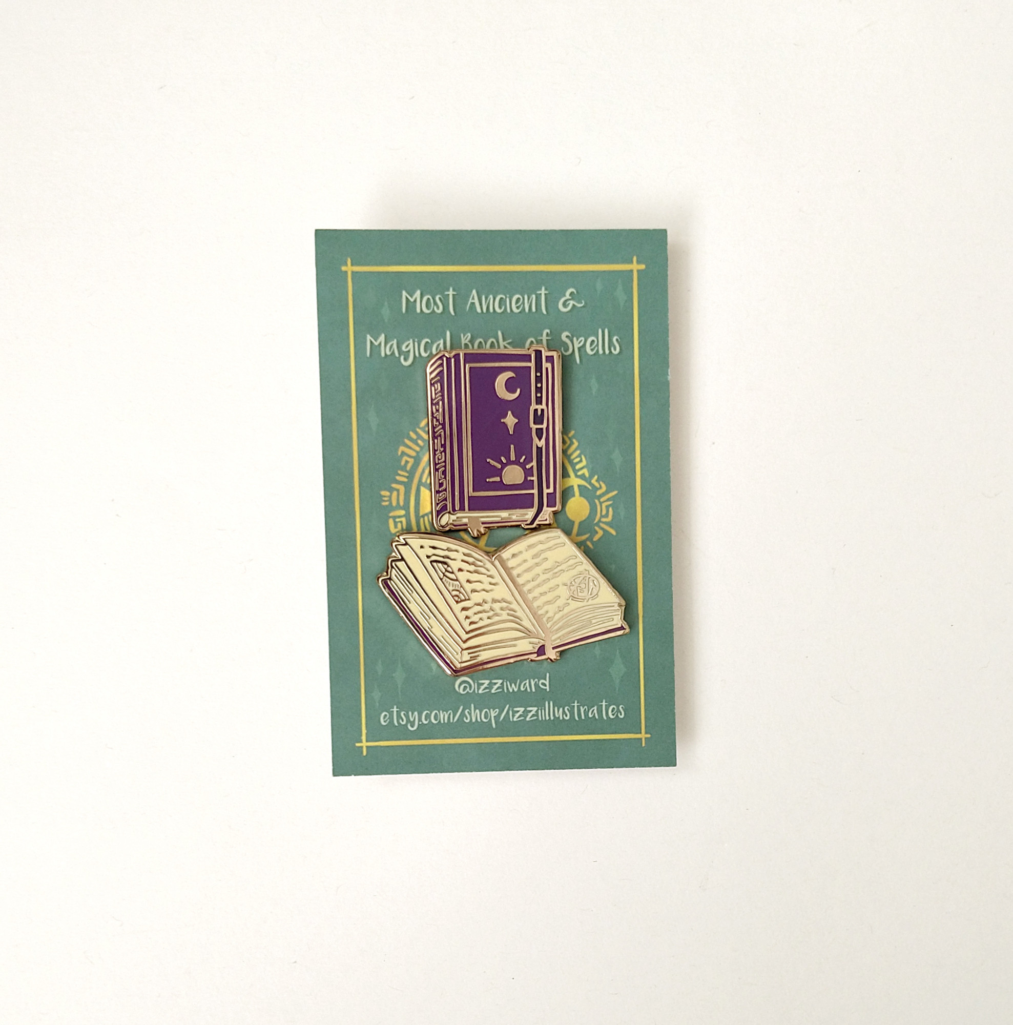 a photograph of two enamel pin badges pinned to a green backing card, against a white background. The pins are shaped like spellbooks, one closed, one open. They are gold metal with purple and white enamel.