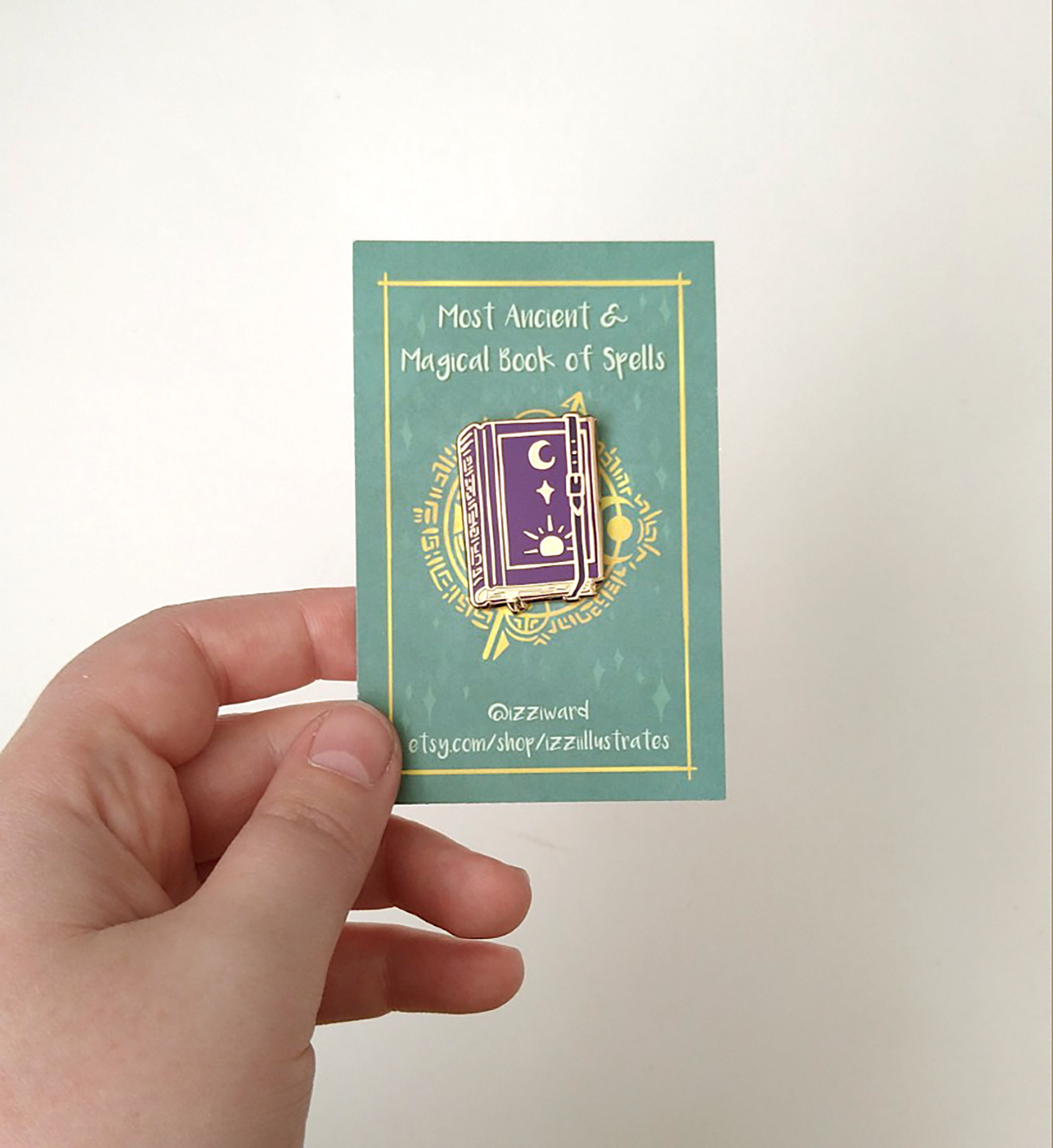 a photograph of an enamel pin badge pinned to a green backing card, against a white background. The pin is shaped like a closed purple spellbook, with gold lines.