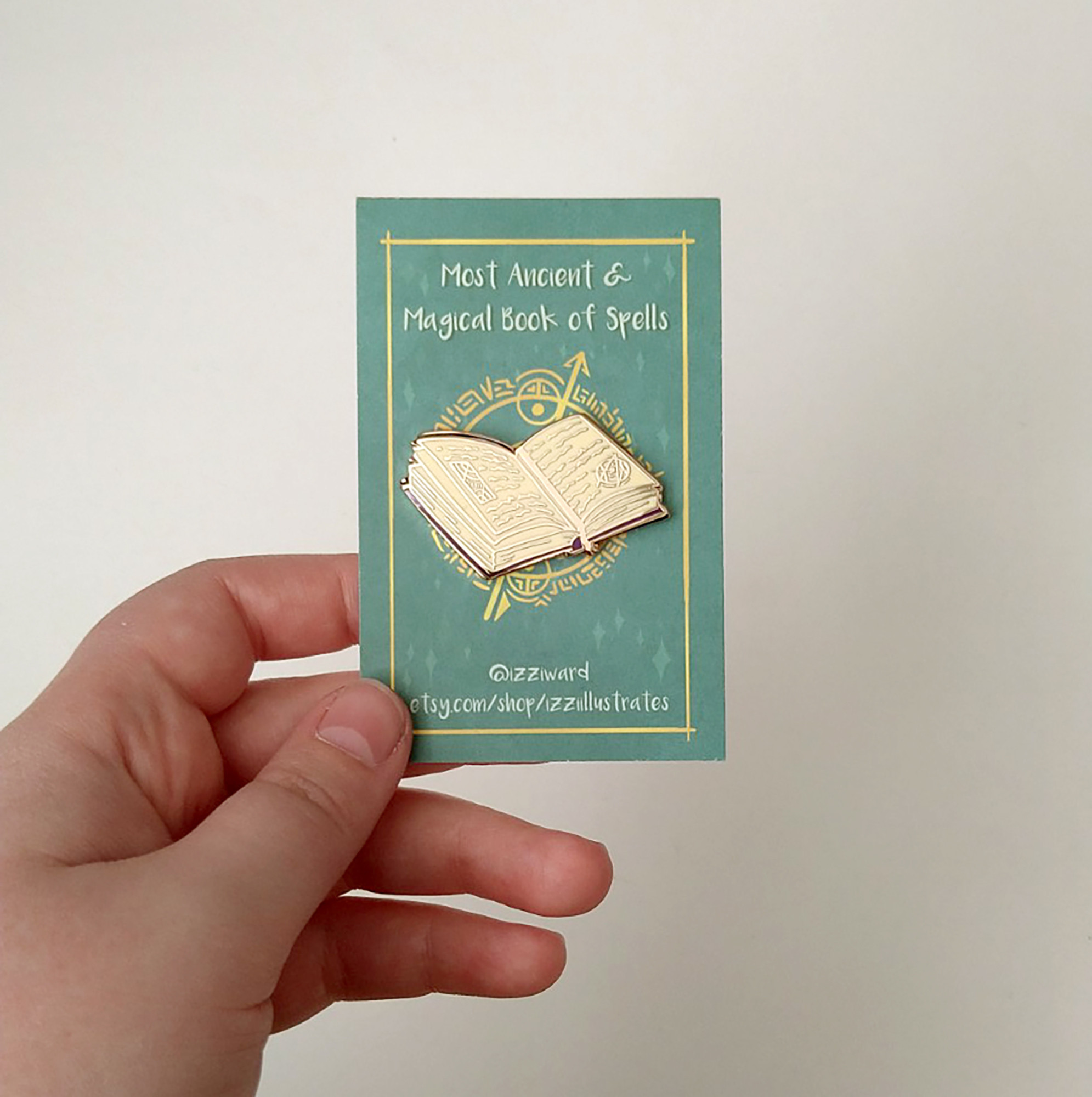 a photograph of an enamel pin badge pinned to a green backing card, against a white background. The pin is shaped like an open spellbook, with gold lines.