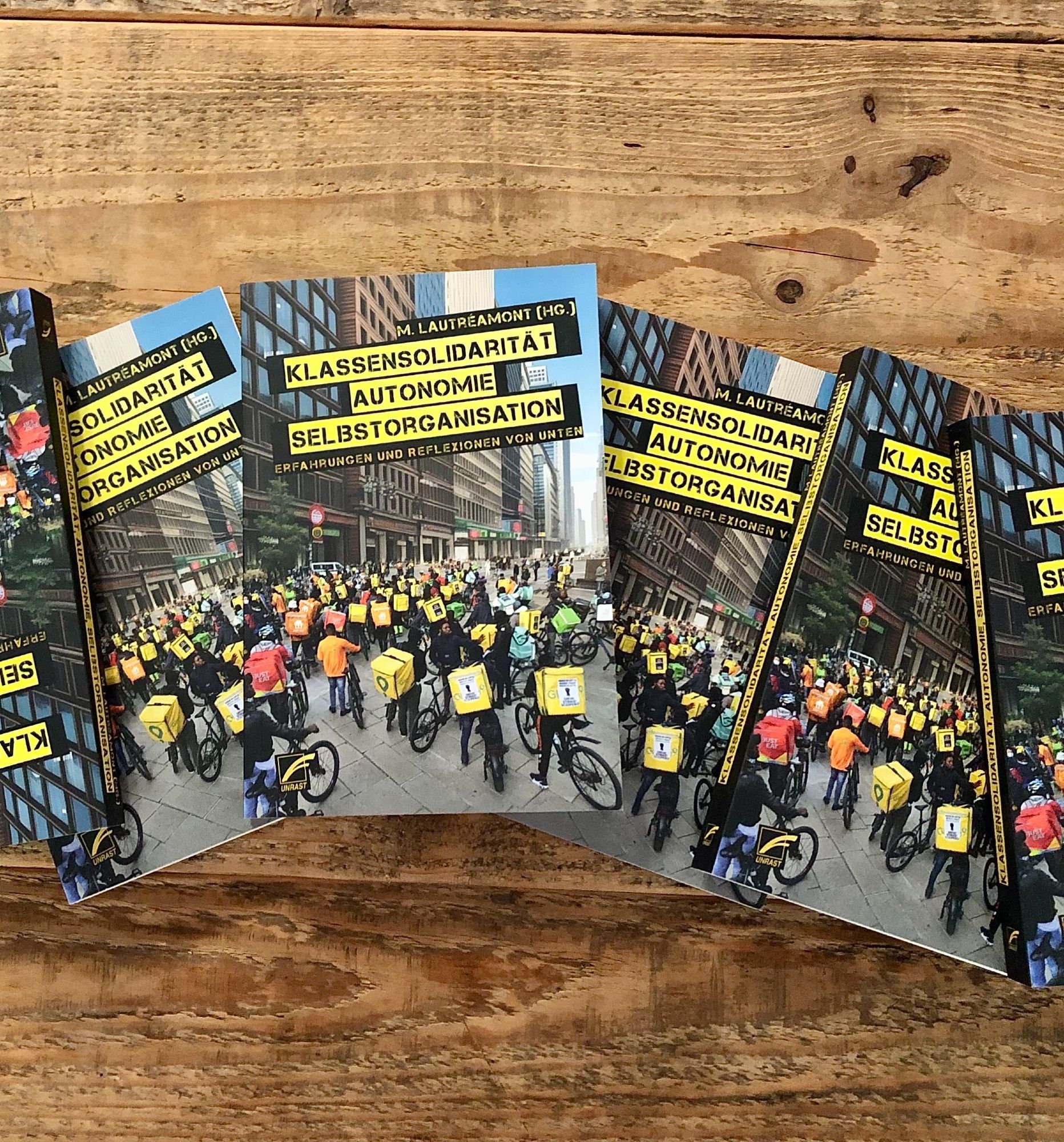 🏷️ Bildbeschreibung: M. Lautréamonts Buch ›Klassensolidarität, Autonomie, Selbstorgaisation - Erfahrungen und Reflexionen von unten‹  liegt mehrmals nebeneinander auf einem Untergrund aus Holz. Mit dem Frontcover zur Kamera. Auf dem Cover ist ein Foto einer Menschenmenge mit Fahrrädern und quadratischen, gelben und orangenen Kurierrucksäcken abgebildet.