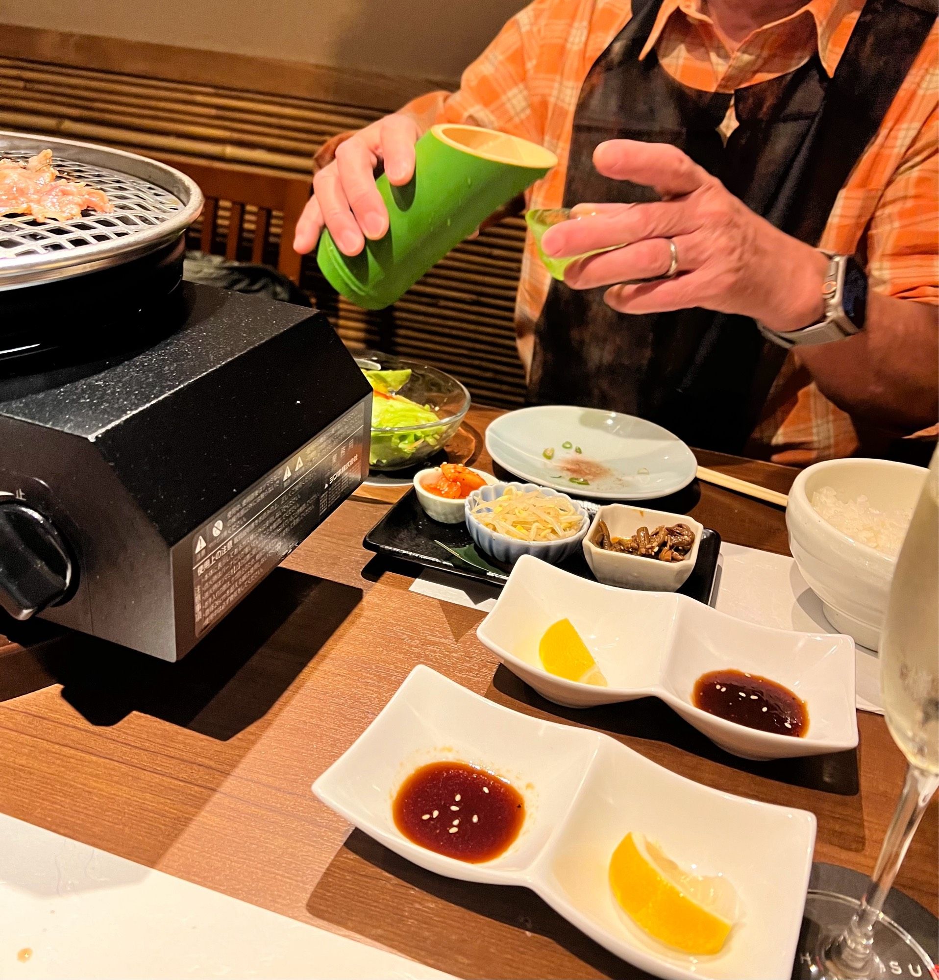 A close-up of hubby pouring sake from a bamboo container at a yakiniku restaurant, surrounded by small dishes and a grill.