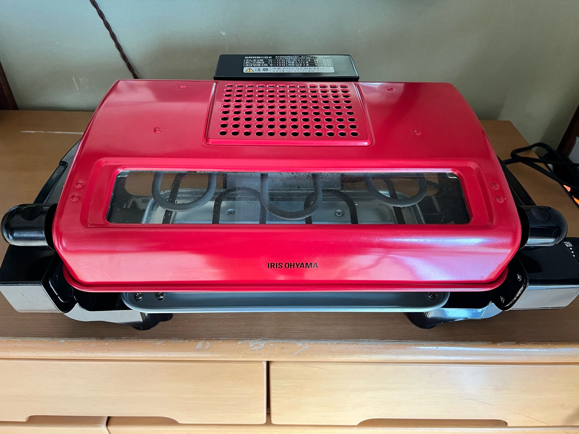 A close-up of a red IRIS OHYAMA fish grill with a transparent window on top, placed on a wooden surface, used to perfectly grill fish.