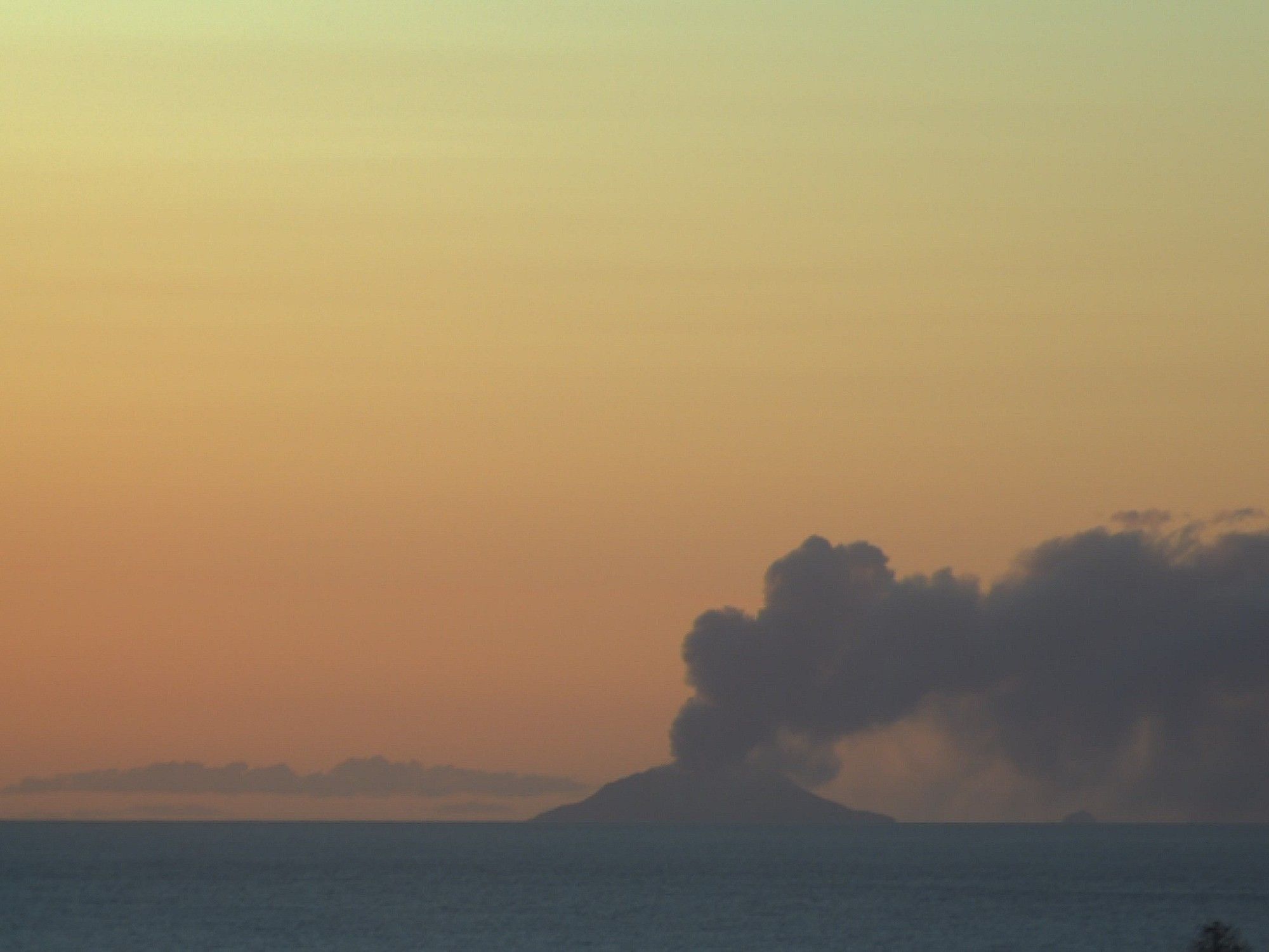 Image of Whakaari from Te Kaha, taken at sunset, with a vividly coloured sky, and a weak eruptive plume