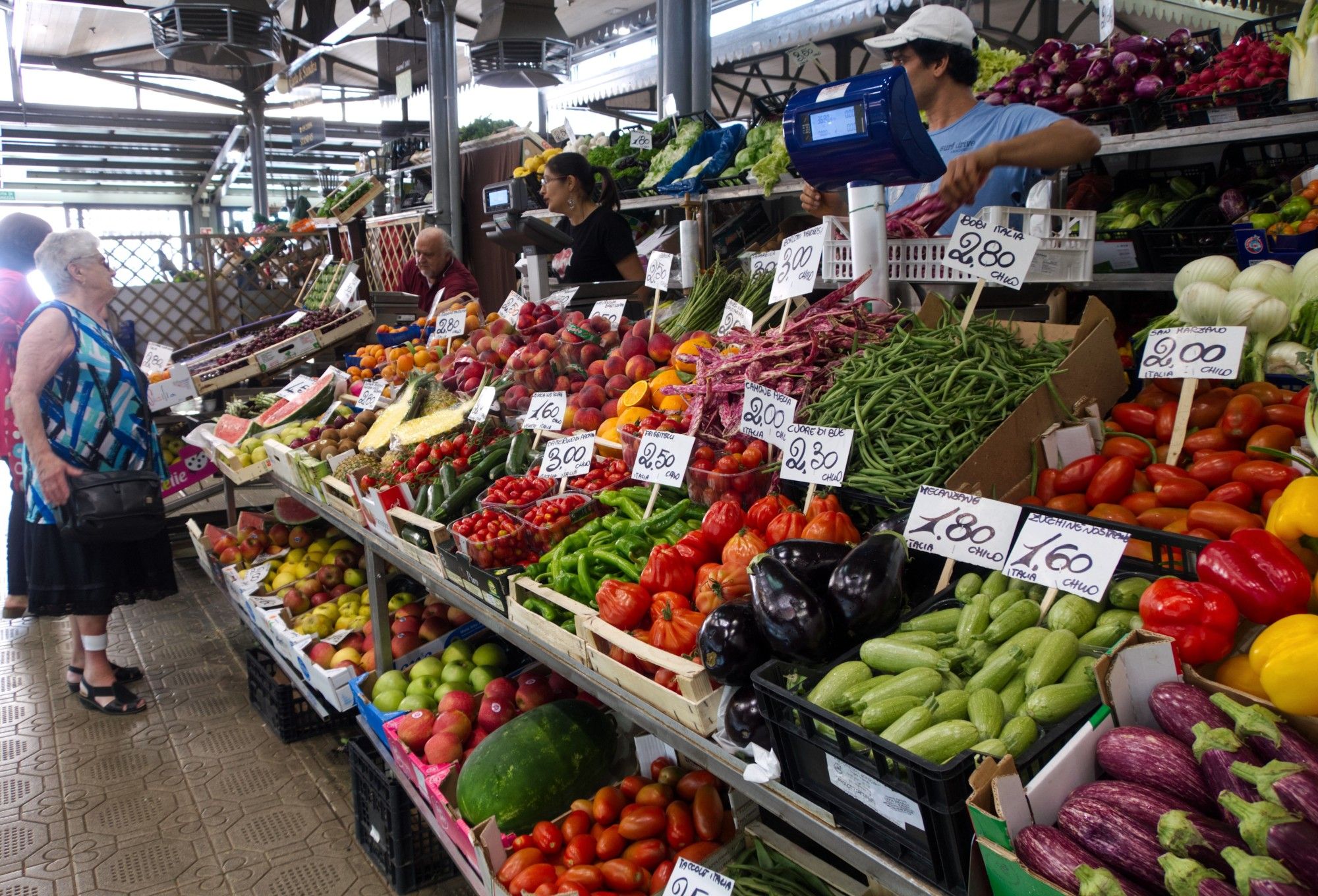 A large fruit and vegetable stand inside a marketplace offering a variety of fruits and vegetables, including watermelon, pineapple, oranges, tomatoes, eggplant, zucchini, etc. all marked with little signs showing the cost (numbers) per kilo. There are 2 ladies on the far left talking to 3 salespeople (1 female and 2 males). *Sorry for the earlier delete… missed 1 salesperson when describing the photo.