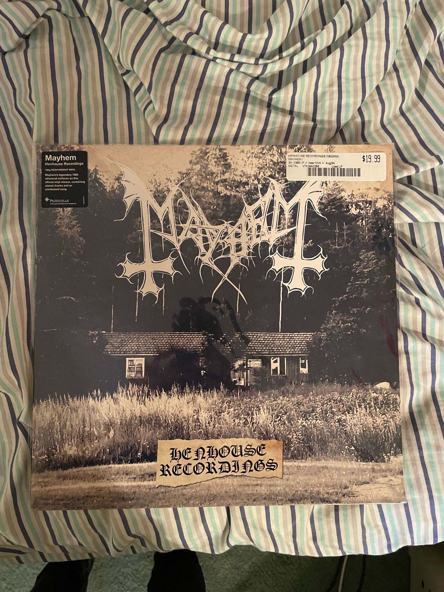 A vinyl copy of the Mayhem record “Henhouse Recordings” sitting on a striped bedspread. It has a typical black metal logo featuring bat wings and upside down crosses and a sepia photo of a farmhouse on it