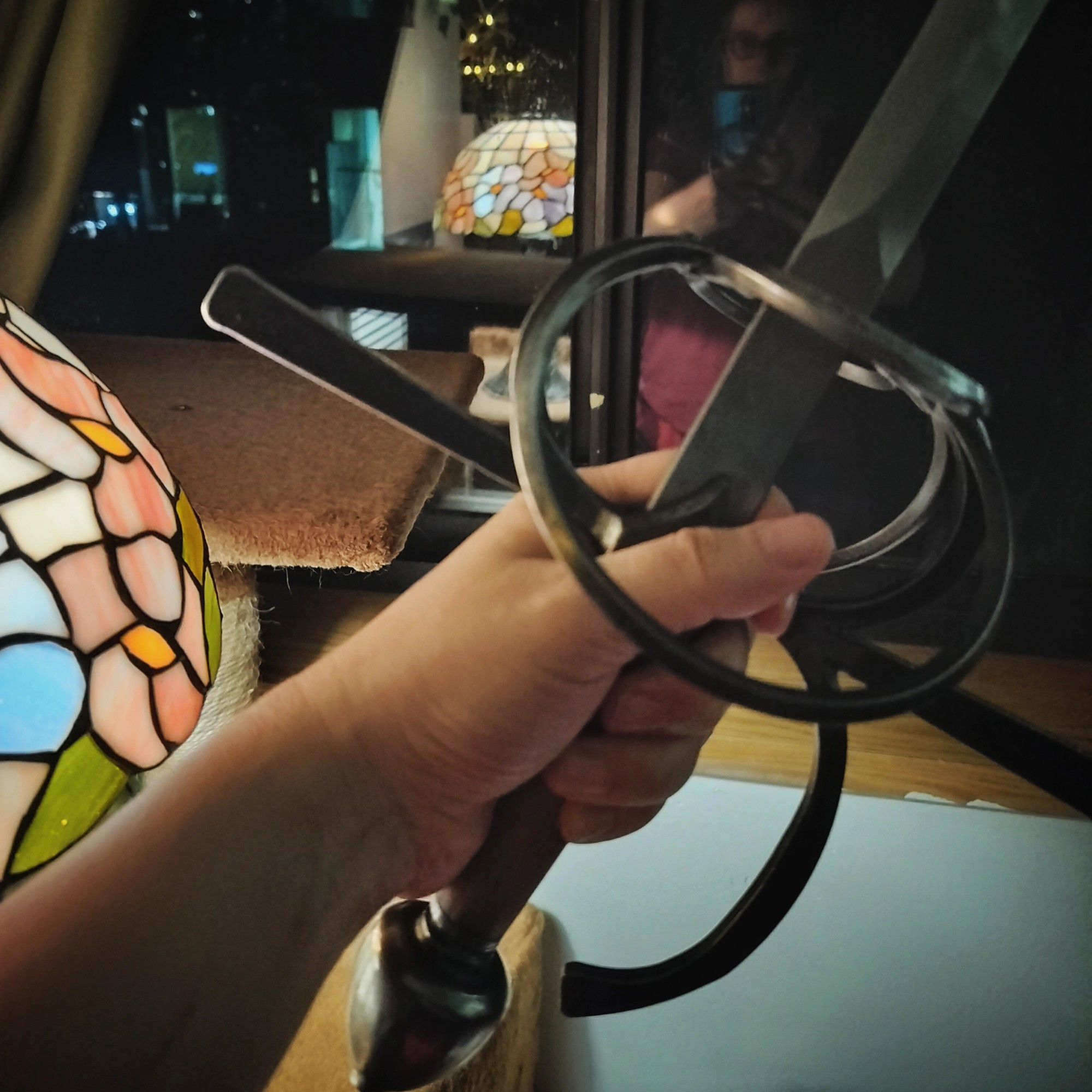 A hand holds a training rapier with a ring hilt. In the background is a stained glass lamp and a window showing nighttime with distant tall buildings. 