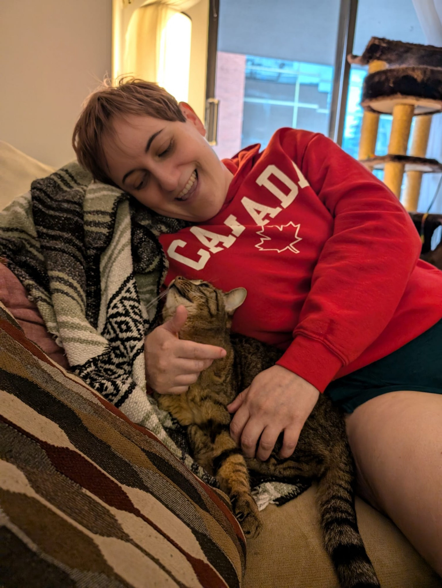 A human is grinning down at a tabby cat that is smiling up at her. They're both sitting on a couch. The human is wearing a red hoodie that says Canada. 