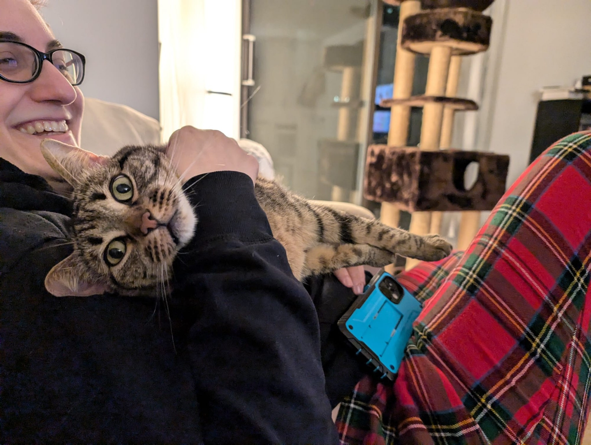 A human wearing plaid pajama pants and glasses is sitting on the couch and holding a cat at a weird sideways angle. The cat is staring very intently at tuff photographer. The human is laughing. 