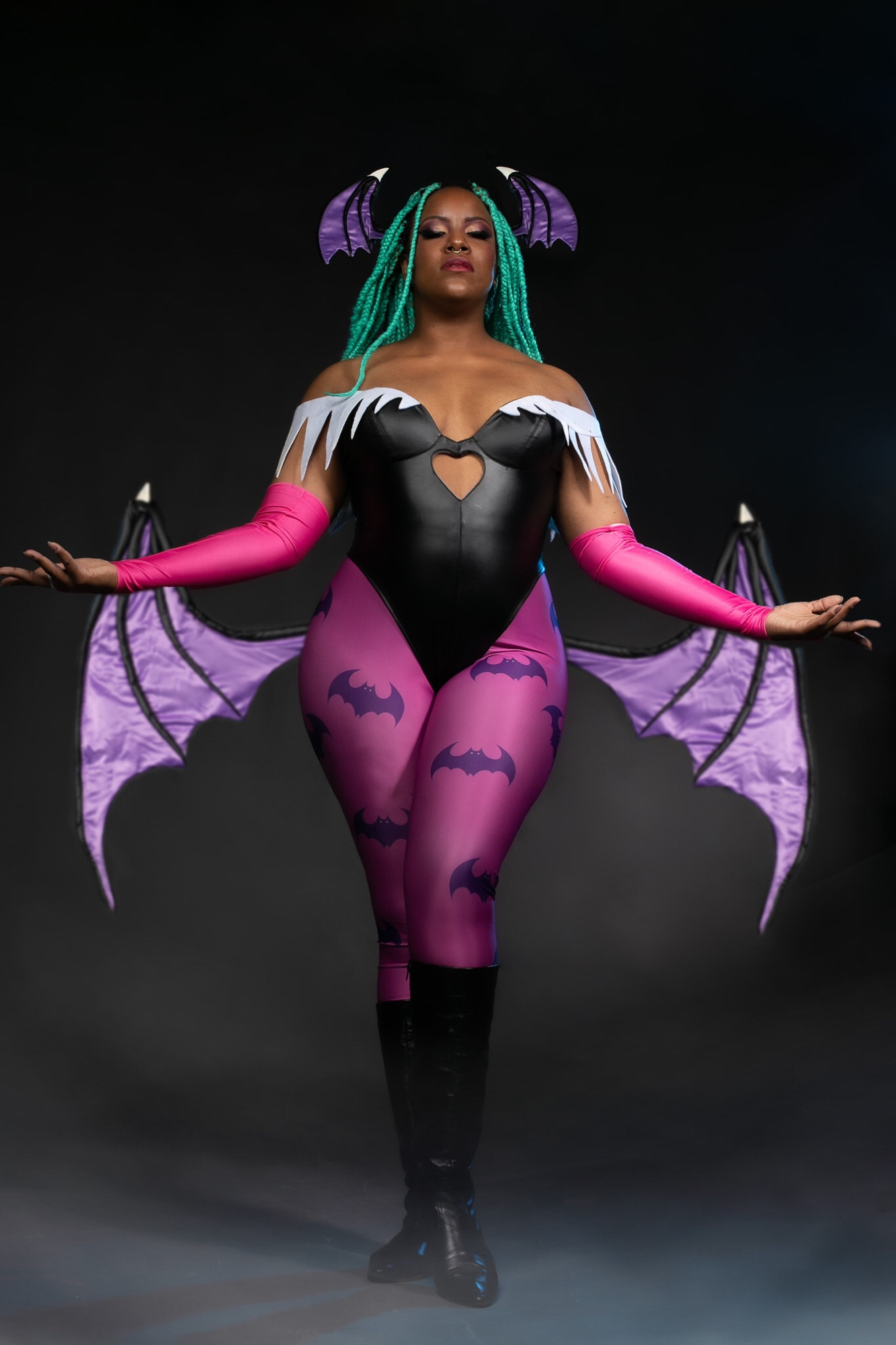 Kami vestindo o cosplay da personagem Morrigan, do jogo Darkstalkers. Morrigan tem cabelo verde/azul turquesa, com asas de morcego nas laterais da cabeça e nas costas. A roupa é um body de courino preto detalhes brancos no decote, com legging pink de estampa de morcegos por baixo, luvas rosa e botas pretas. A cosplayer está posando com os olhos fechados, com os braços suspensos nas laterais do corpo.