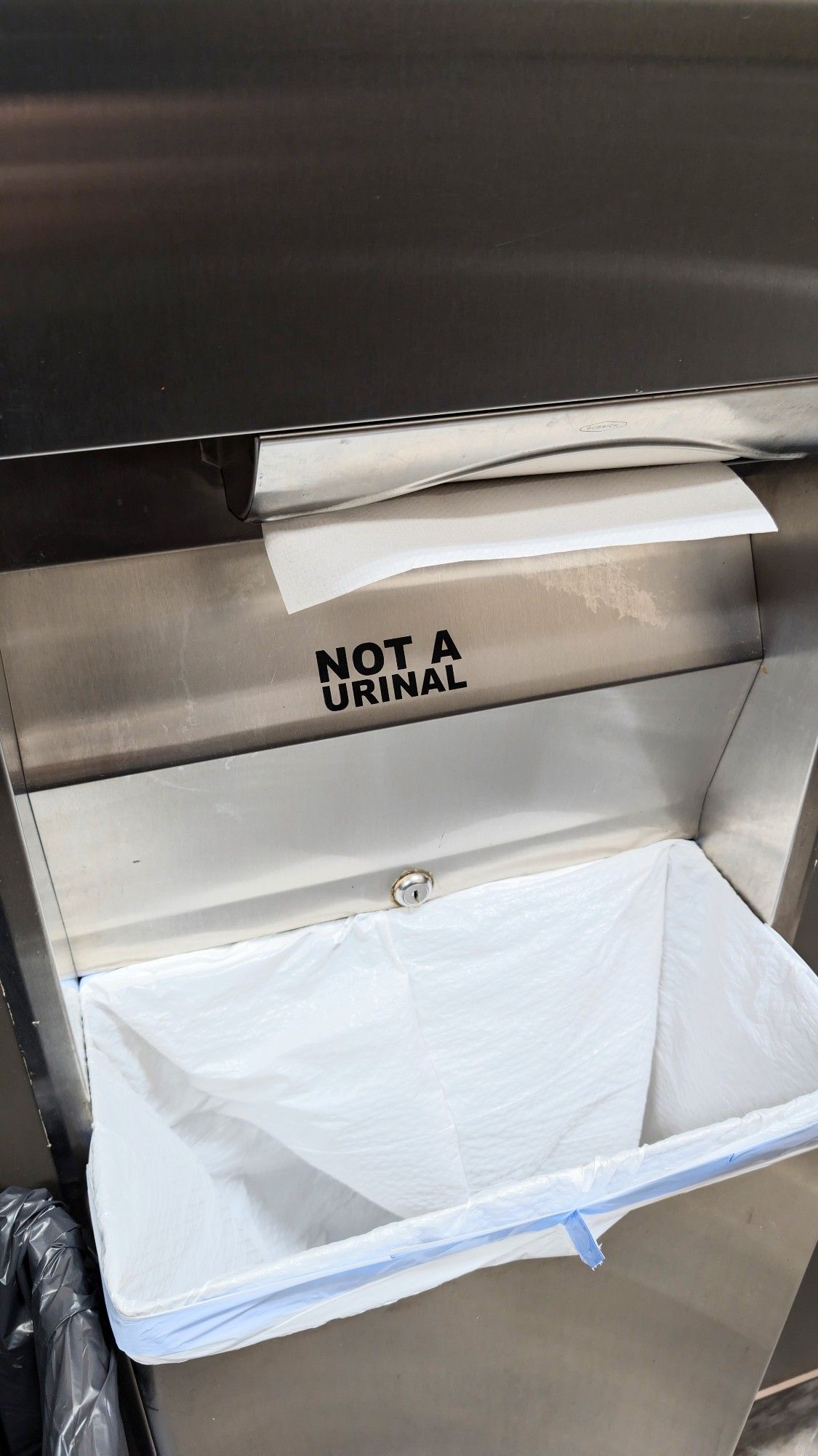 Picture of a paper towel dispenser with garbage can below. Someone put a sign on it that says, "Not a urinal".