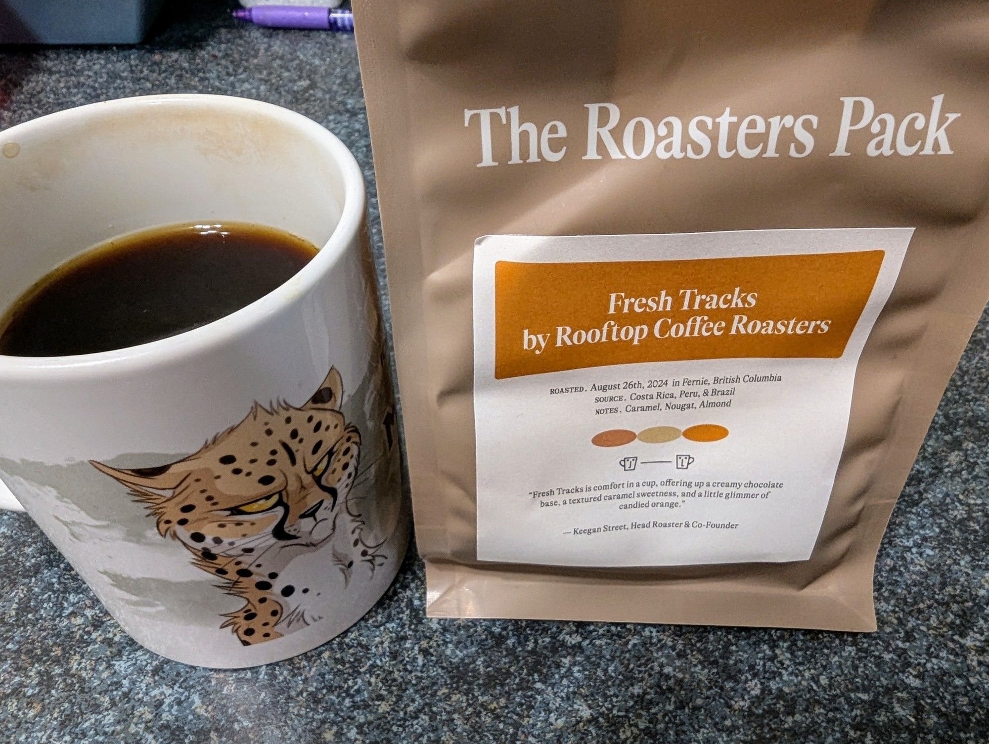 A bag of coffee from Roasters Pack. The coffee is called Fresh Tracks by Rooftop Coffee Roasters in Fernie, BC. The coffee is a blend from Costa Rica, Peru, & Brazil. The tasting notes provided are caramel, nougat, and almond. Next to the bag of coffee is a mug with a cartoon grumpy cheetah.