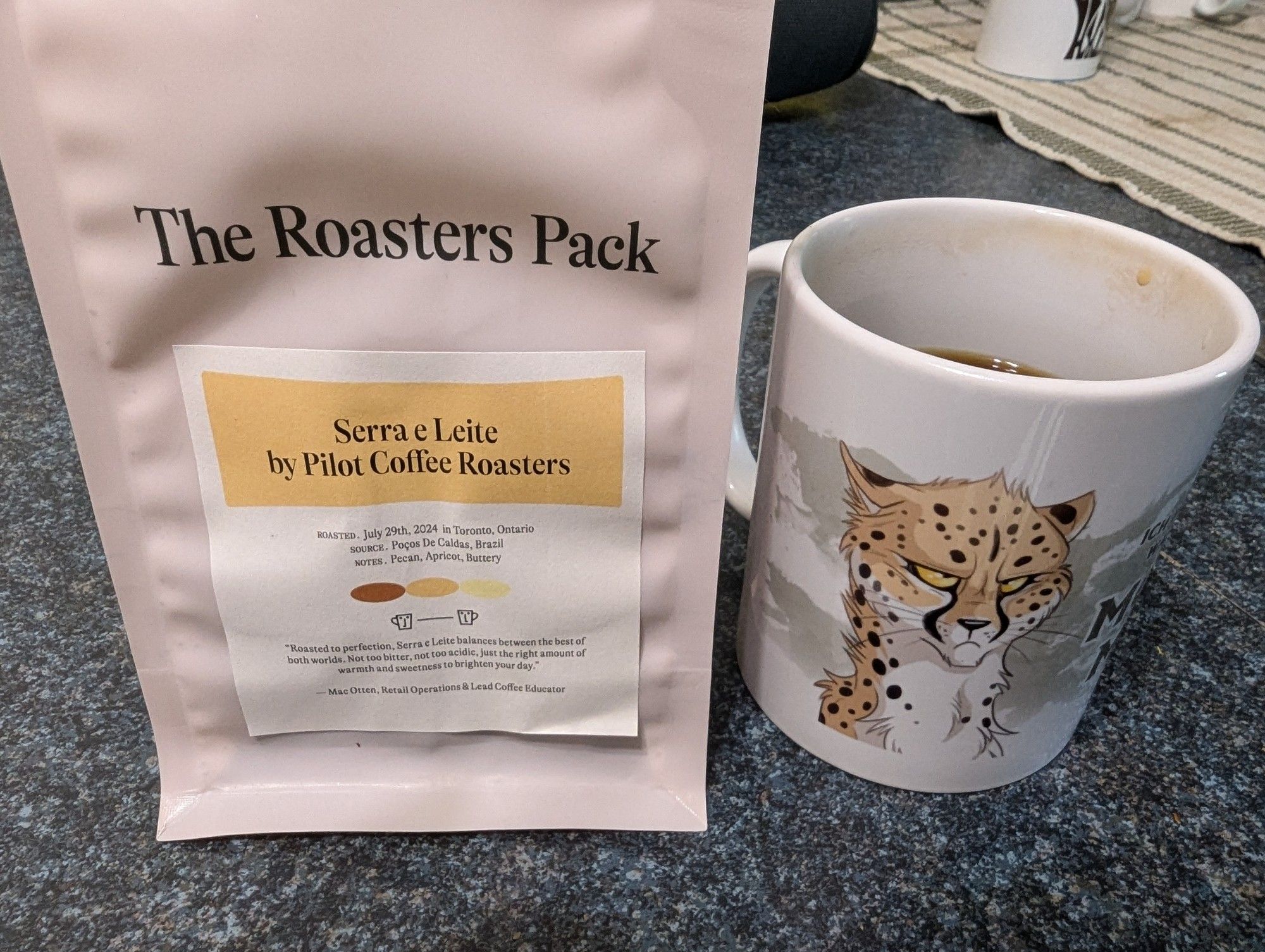 A bag of coffee from my Roasters Pack. The coffee is called Serra e Leite by Pilot Coffee Roasters in Toronto. The coffee's source is Poços De Caldas, Brazil. The tasting notes provided are pecan, apricot, and buttery. Next to the bag is my mug with a drawing of a grumpy cheetah.