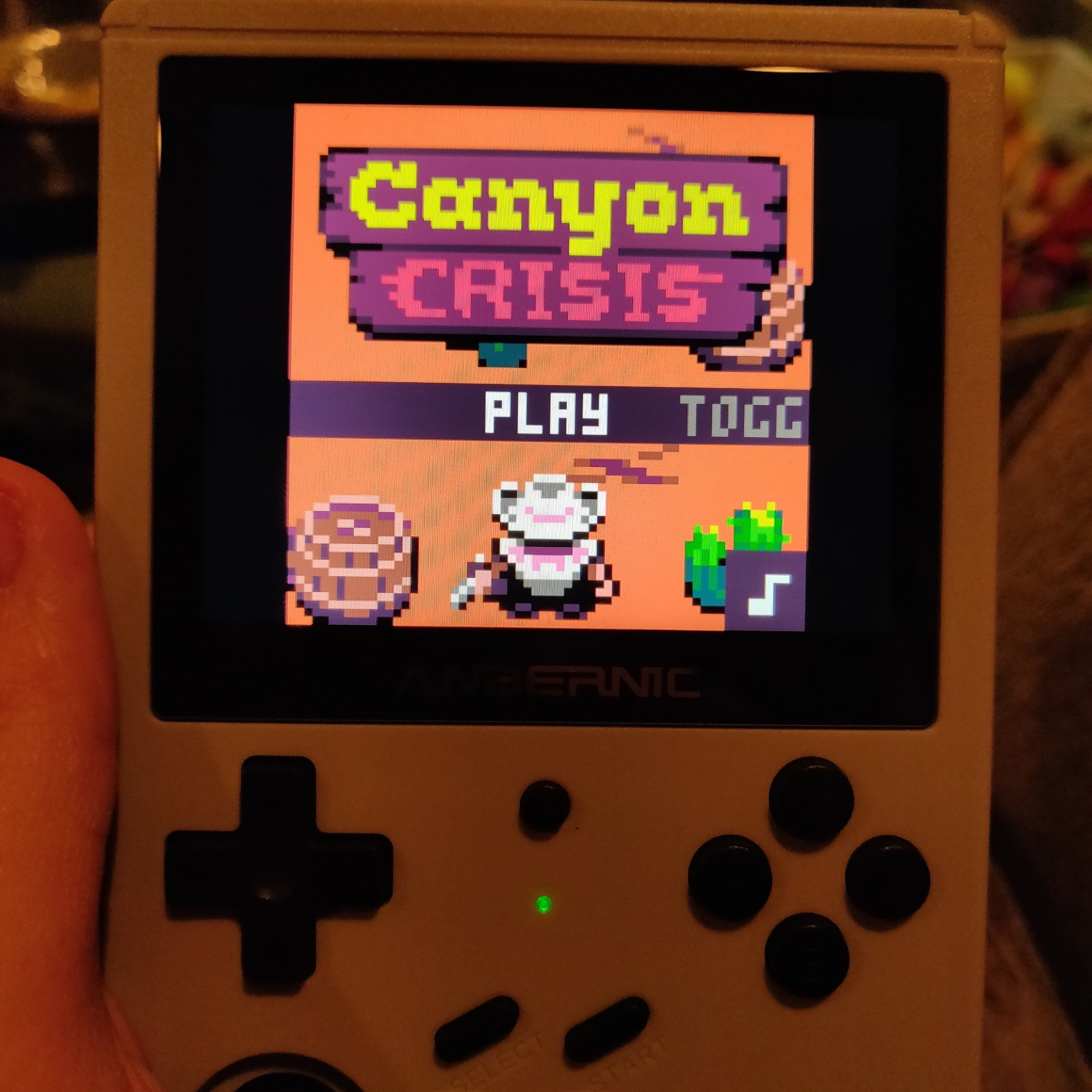 A picture of a hand holding a small gaming device, called an Anbernic RG351V. On the device's screen is the main menu from Canyon Crisis.