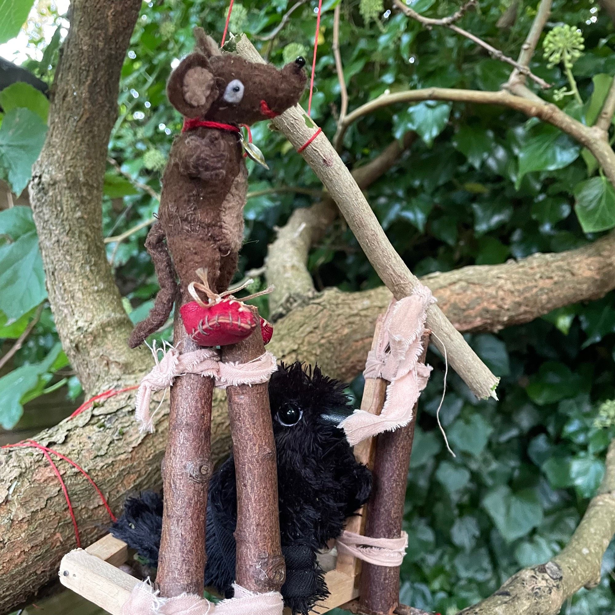 Photo of Silvius the little Latin mouse and Dusky the young crow, working on their tree house. This time, they have fixed logs upright by the door opening, tying them in place with thin cloth strips. Now they are lifting another log to be the lintel. Dusky is holding one end in place with her beak, while Silvius lowers the log with a red string pulley