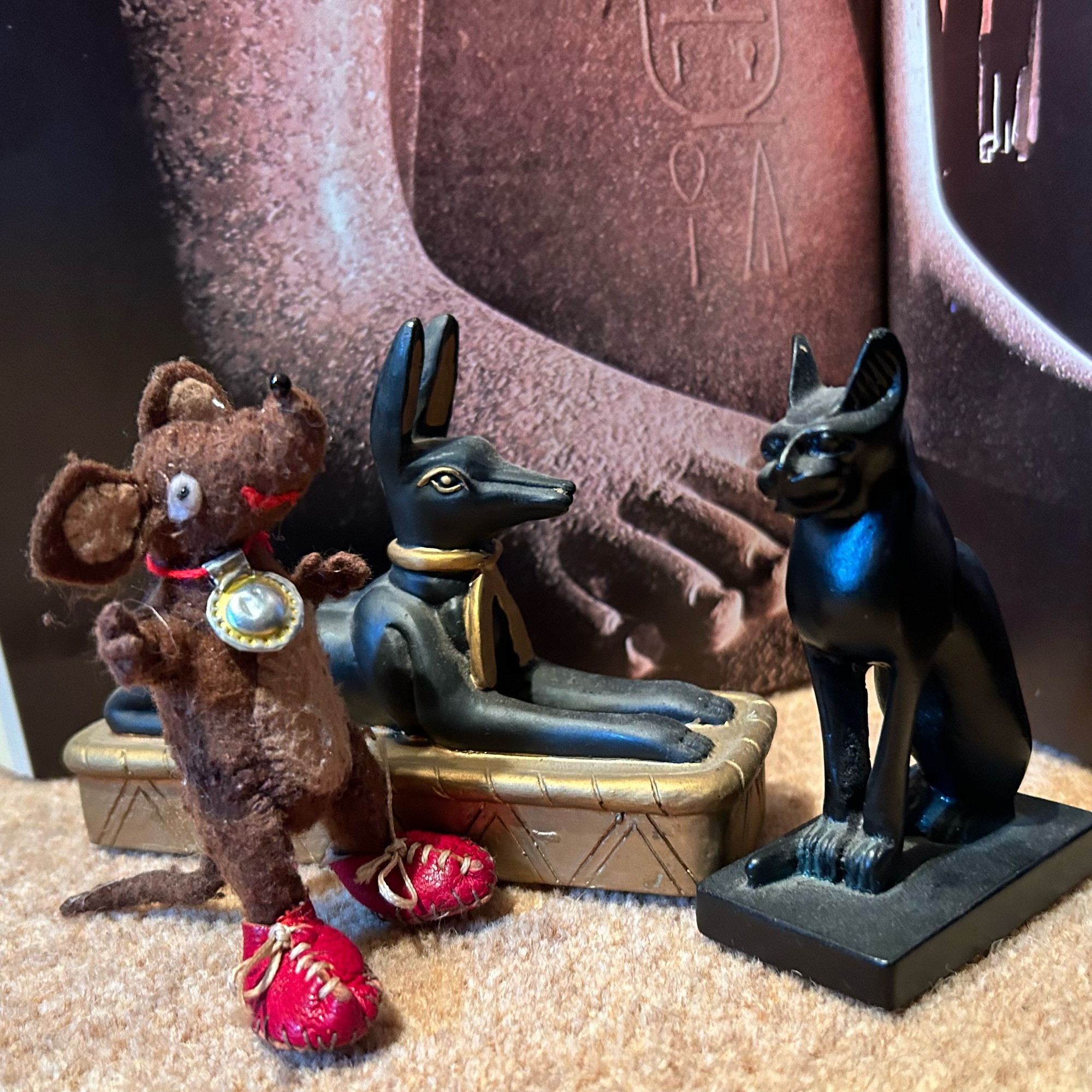 Photo of Silvius, the little Latin mouse, with a backdrop of a massive Egyptian statue. He's looking at little black stone statuettes of Ancient Egyptian gods, still recognised and worshipped in the Roman province of Egypt even in Silvius' time. 