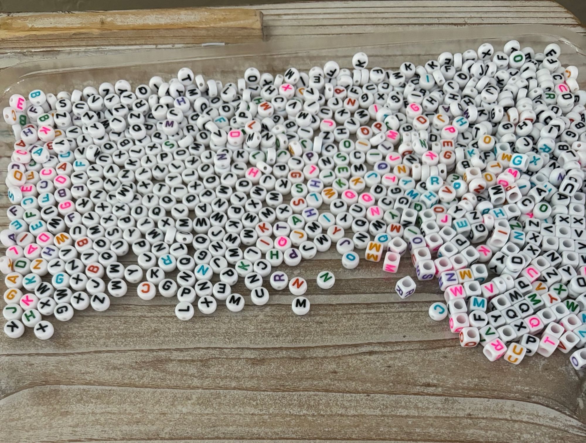 spilled letter beads: circular, cubes, black, and colorful. not sorted, just a mess of beads of a light wood tray.