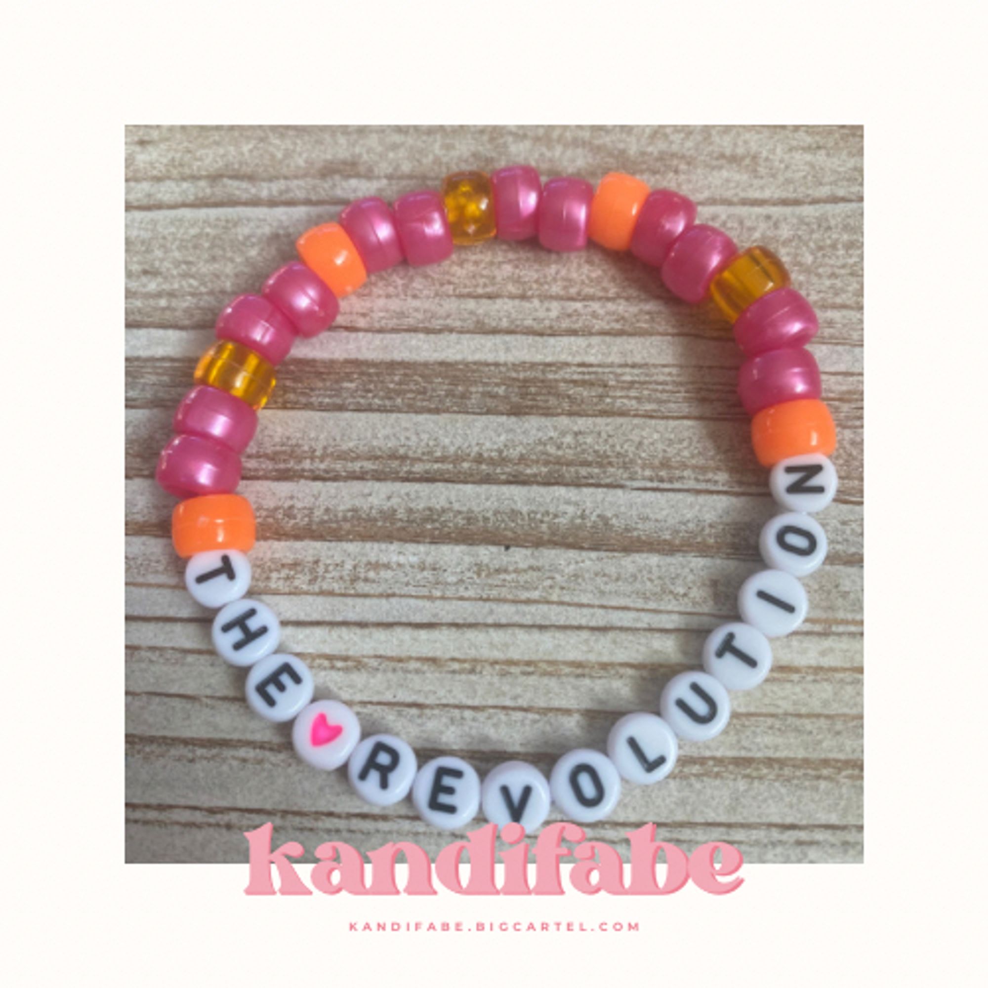 A kandi bracelet that reads “THE (pink heart) REVOLUTION” in black letters on white circle beads. The rest of the bracelet is filled in with a pattern of opaque and translucent orange and pearlescent pink pony beads.