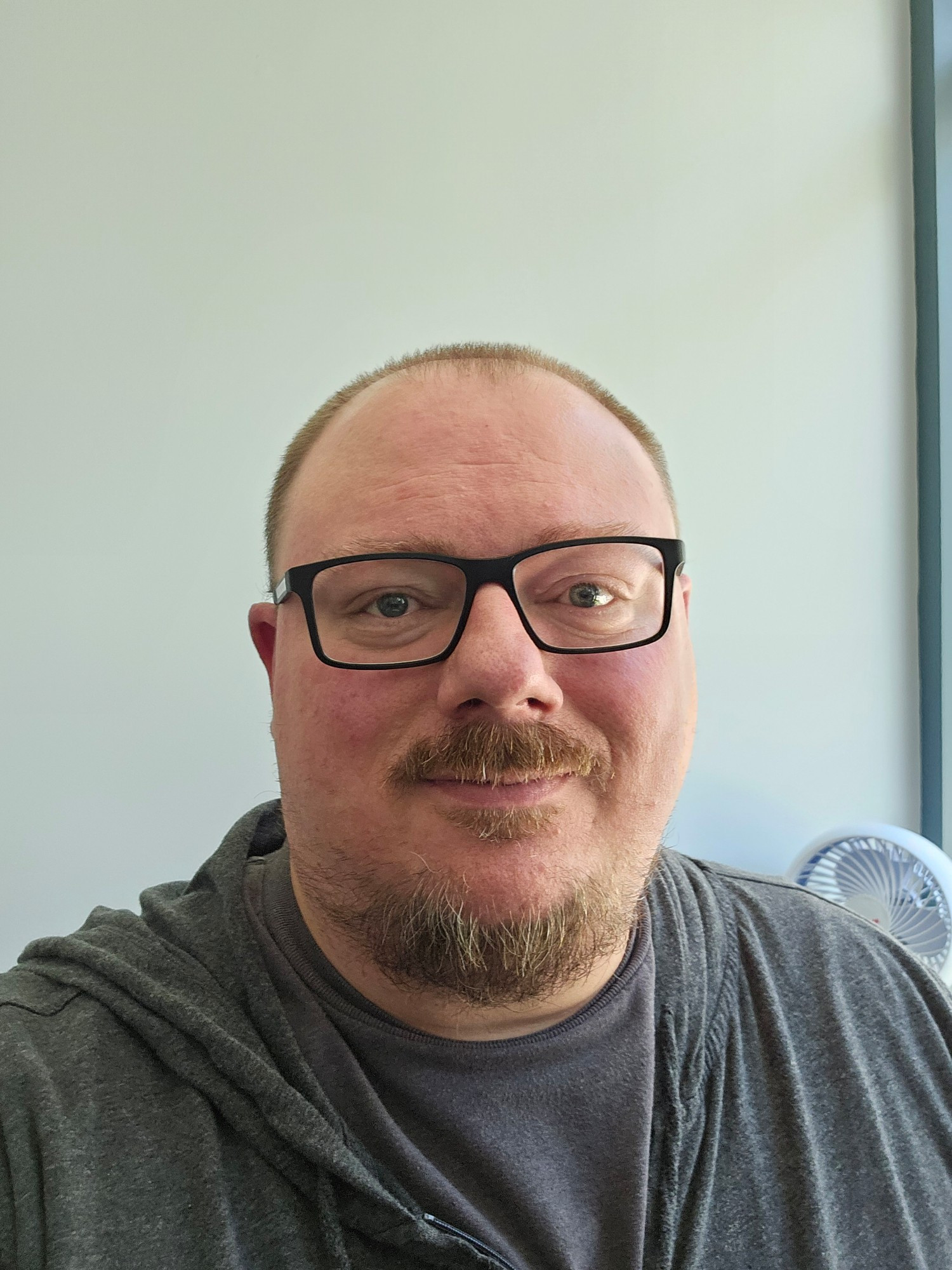 40 year old white man with short cut hair, a mustache, a spotty beard, and square black glasses. He's wearing a gray shirt and gray hoodie.