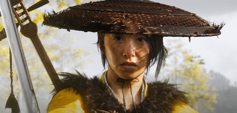 Screenshot from the GHOST OF YOTEI trailer, depicting an asian woman (portrayed by Erika Ishii) in a yellow, fur-lined robe and tattered straw hat, holding a sword at the ready, with a shamisen (I think?) on her back. She's looking past the camera tentatively, as if unsure of what an opponent is going to do.