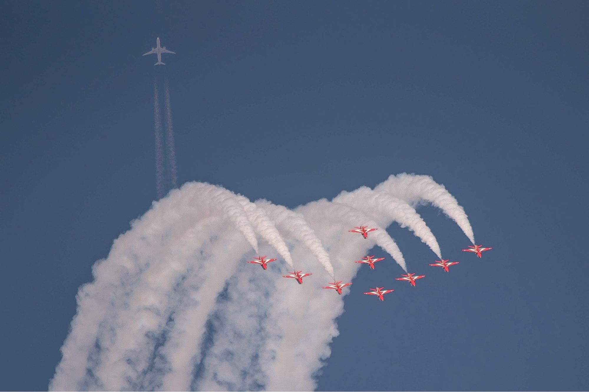 The Royal Air Force’s Red Arrows get photobombed by a Boeing 787 Dreamliner flying high overhead as the demonstration team performs over London, Ontario, Canada during Airshow London 2024.