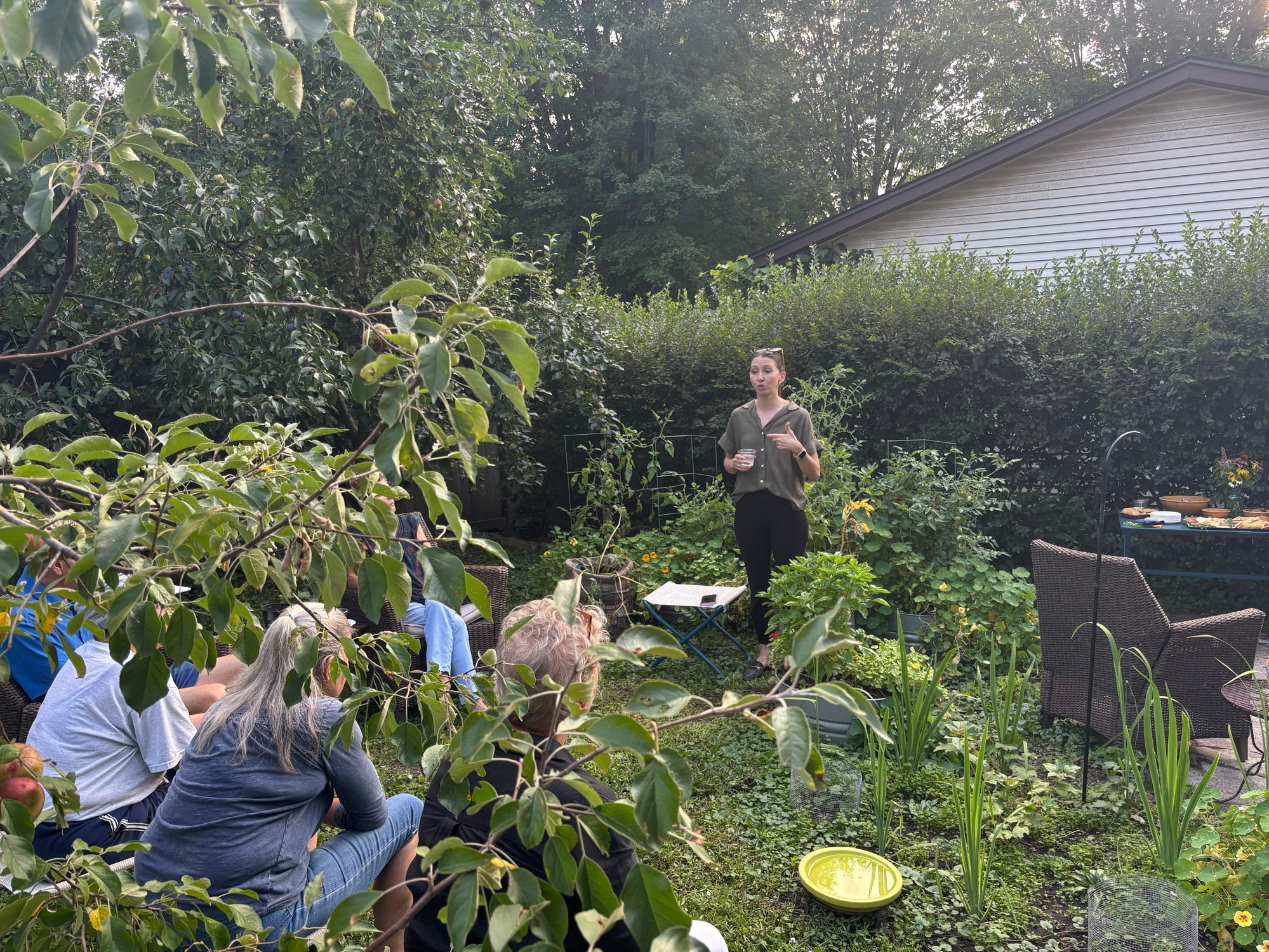 Moly standing talking to a group of seated neighbors in a garden.