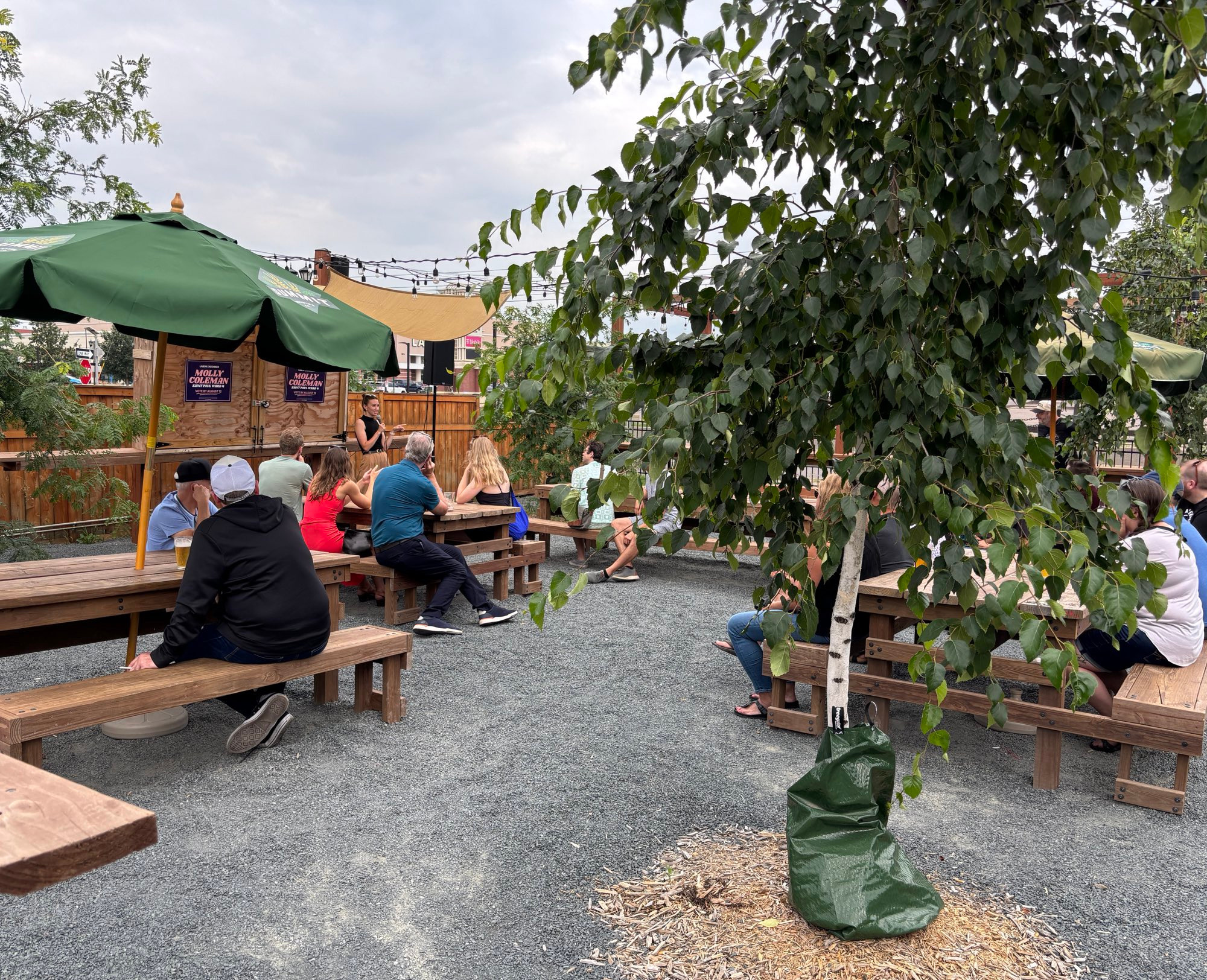 Molly talking into a microphone on a bar patio while people listen at wooden picnic tables 