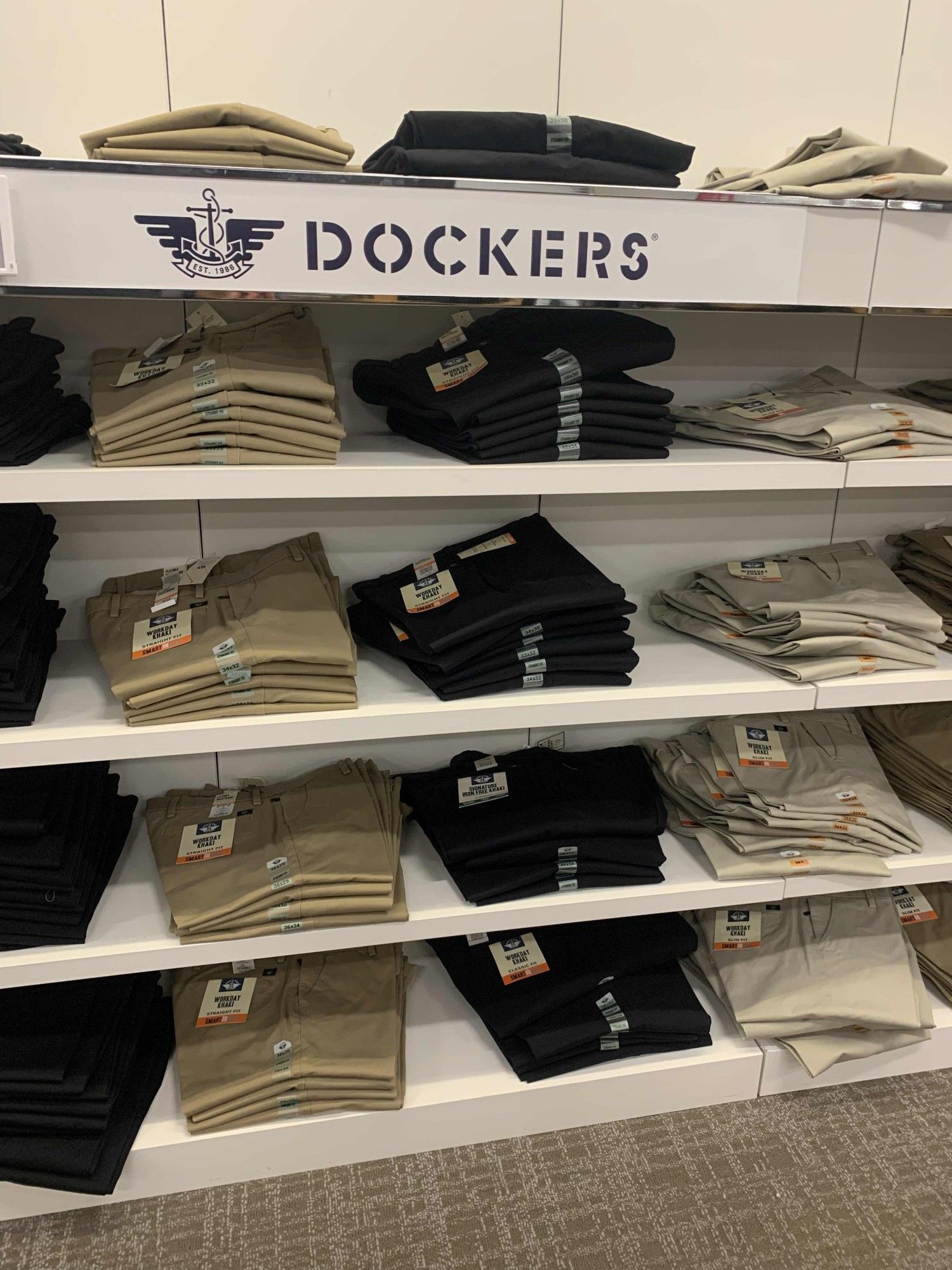 a selection of Dockers brand pants on five white shelves. from right to left the colors of the Dockers are as follows: black, khaki, navy blue, and sand.