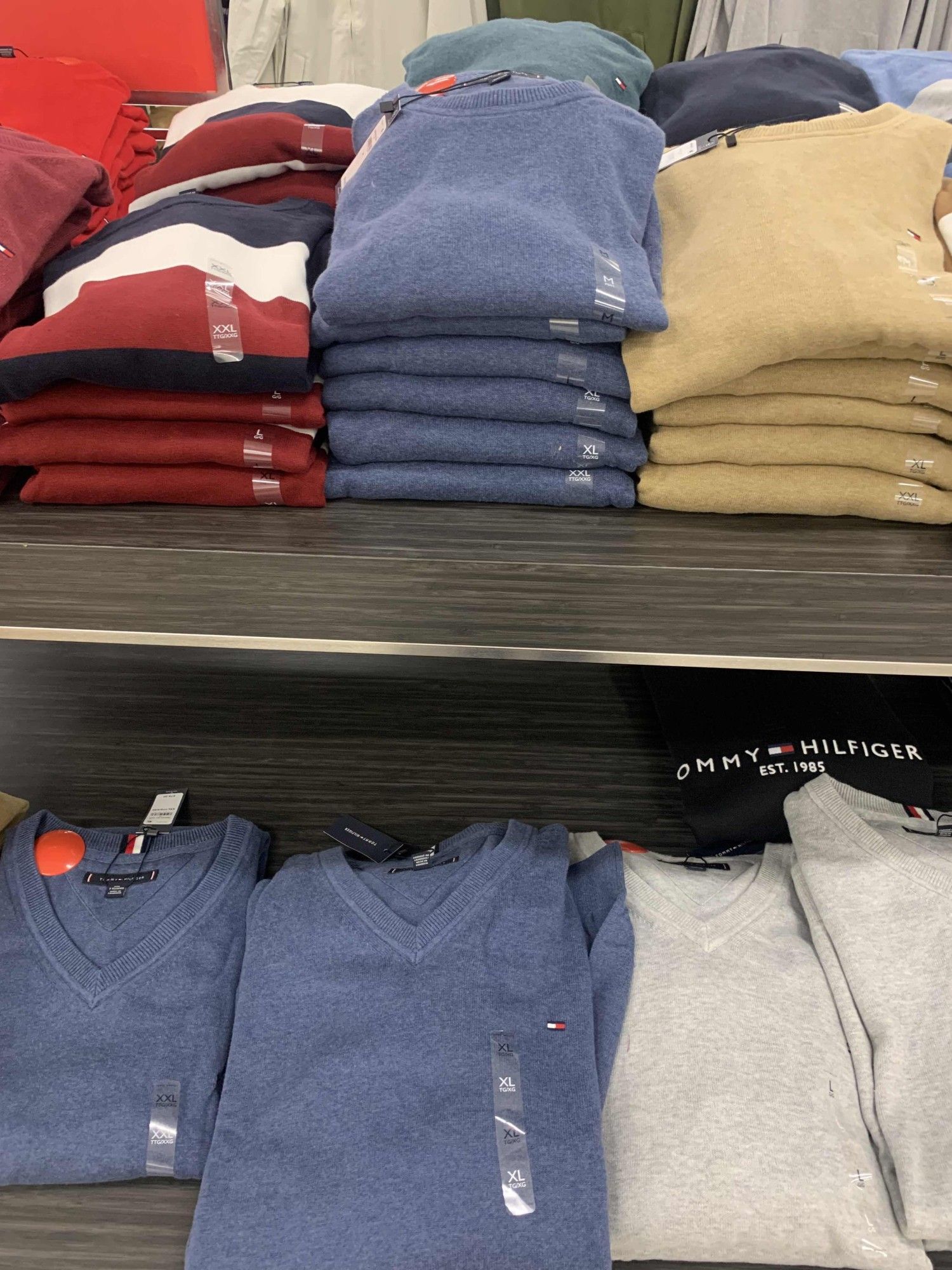 two shelves of colorful v-neck sweaters. the top shelf from left to right holds: red, white, and blue sweaters, a baby blue pile, and yellow pile.  the bottom shelf is more the same, though two stacks of baby blue sweaters, and two of light gray.