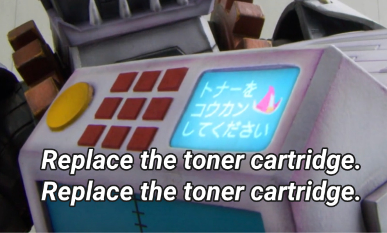 Cropped Screenshot of Zenkaiger, showing a copy machine with an error in its screen, with a little pink hat, similar of Zenkai Magine's. It also has subtitles, which read "Replace the toner cartridge. Replace the toner cartridge"