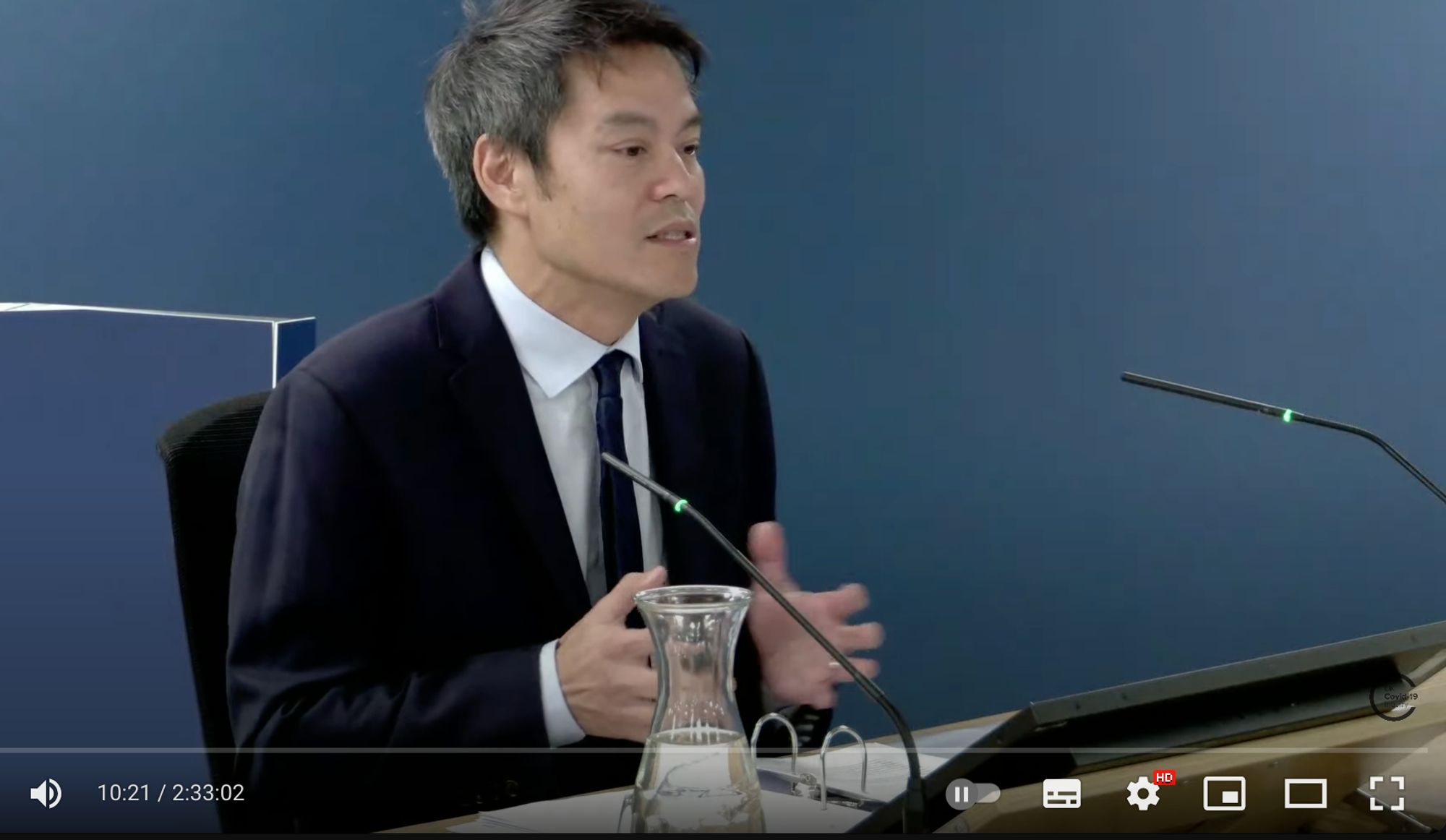 Screenshot aus YouTube Video: Professor Fong sitzt an einem Redepult mit 2 Mikrofonen und erläutert die Lage.