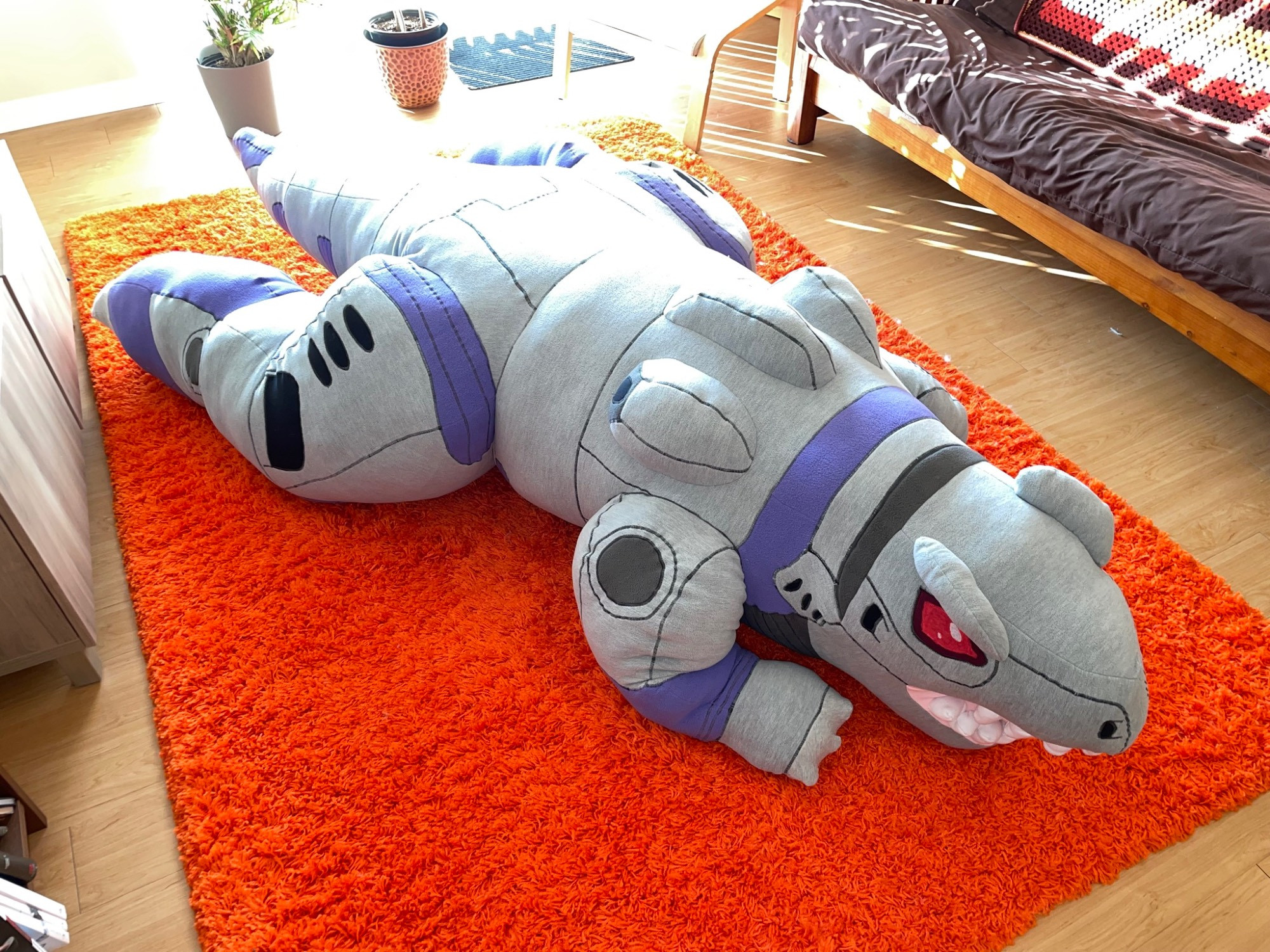 A photograph of an overstuffed half scale plush of Zeke the organoid from Zoids Chaotic Century on an orange carpet.  It’s a grey and purple theropod dinosaur robot and it will guard your dreams at the cost of causing a bed gravity singularity.