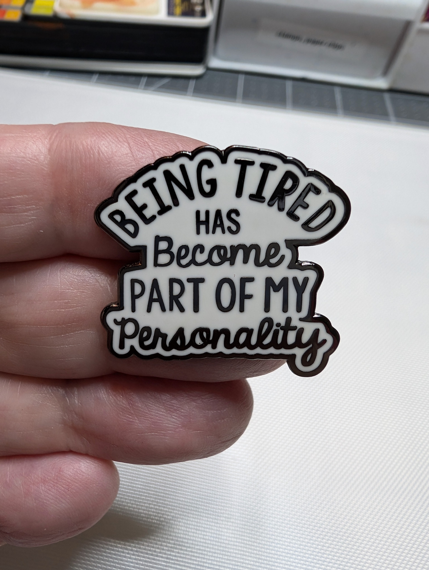 close up of an enamel pin being held in my hand. Black text on white background. Text reads: "Being tired has become part of my personality"