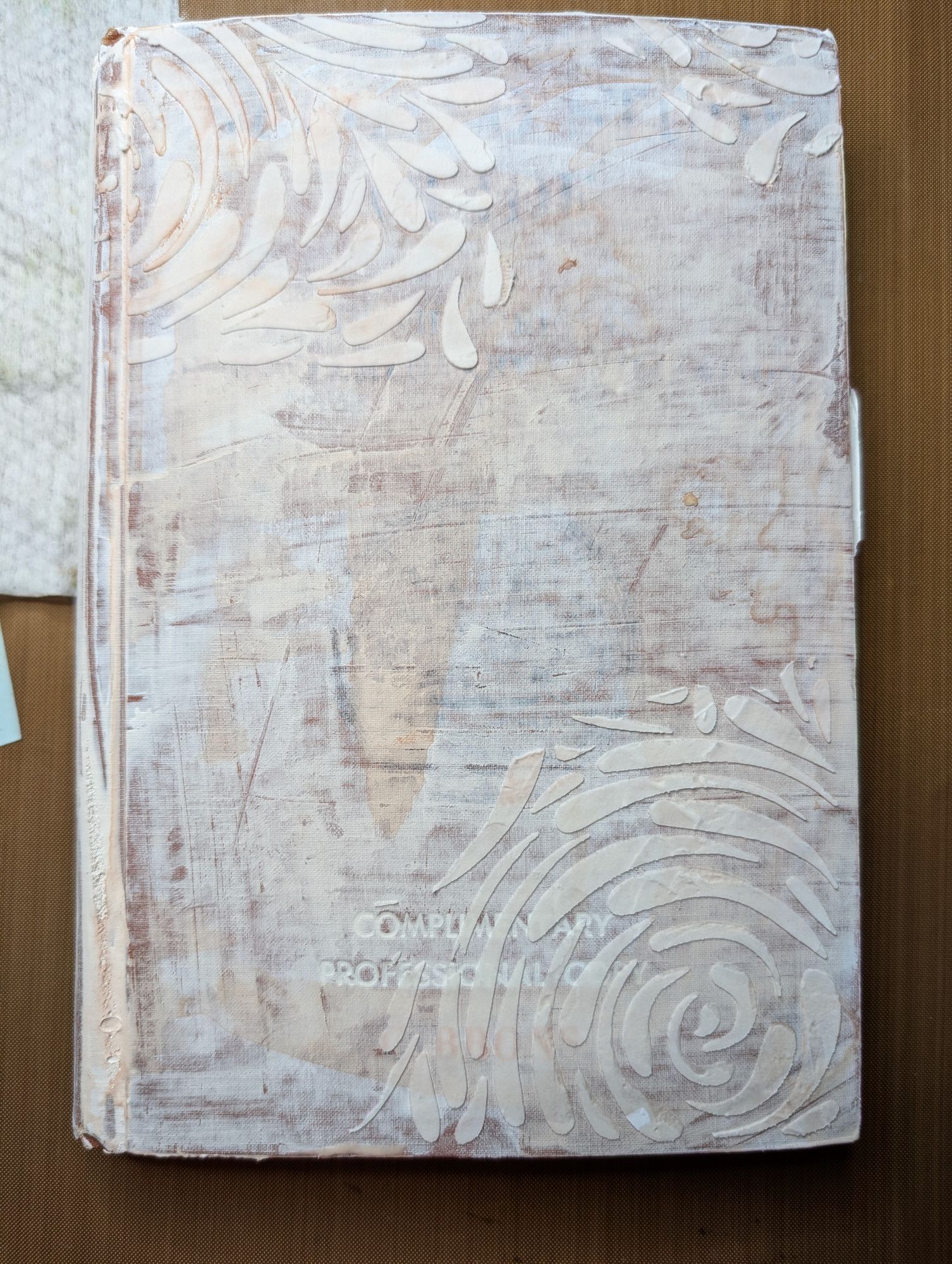 photo of the same book, but the cover is now painted in white gesso & there is a swirly floral stencil pattern in white texture paste in the upper left & lower right corners.