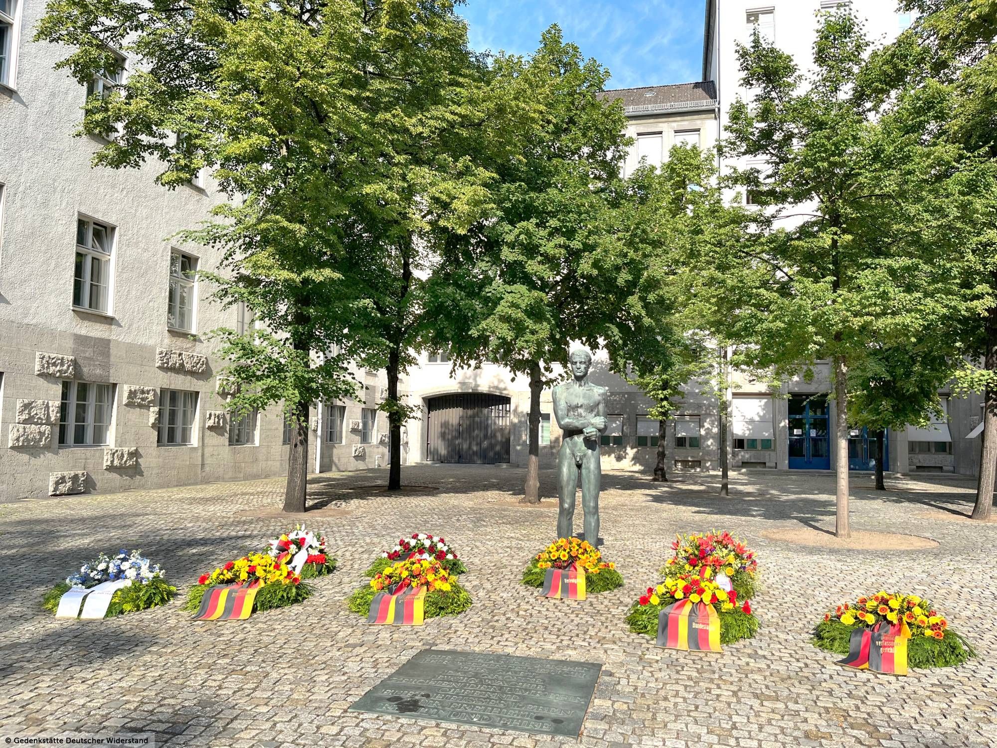 Das Foto zeigt den Ehrenhof der Gedenkstätte Deutscher Widerstand mit der Skulptur des “Gefesselten Jünglings” zwischen sechs Bäumen und einer Bronzeplatte mit Inschrift, die in den Pflasterboden eingelassen ist. Davor liegen neun Kränze mit Blumenbouquets und Kranzschleifen in den Farben Schwarz-Rot-Gold