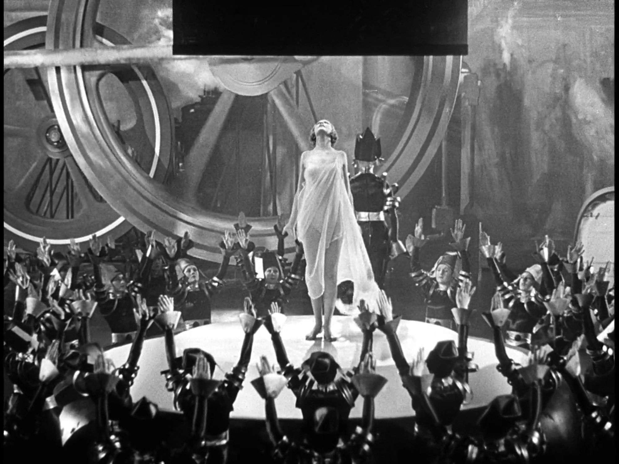 Evergreen (1934). On a small circular stage, a woman in a sheer white shift dress looks up as a piston comes down on her to transform her into a weapon of war. The previous woman who underwent this is walking away, dressed in futuristic black-and-silver armor. The stage is surrounded by women dressed in the same armor, lifting their arms in exultation of the war machine. Behind them all, giant industrial flywheels