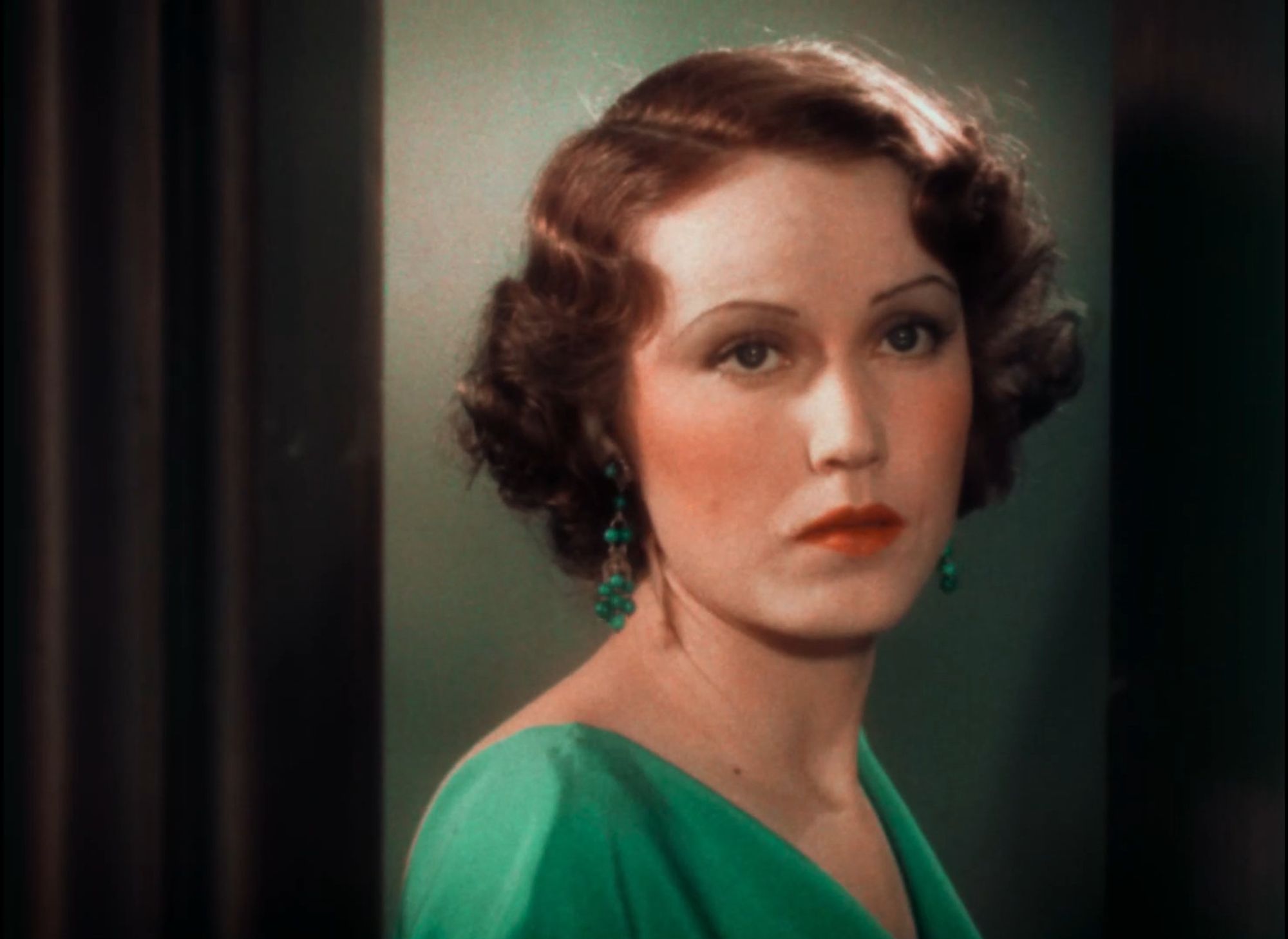 Still of a woman facing but looking past the camera. Short brown hair with marcel waves, emerald green blouse.