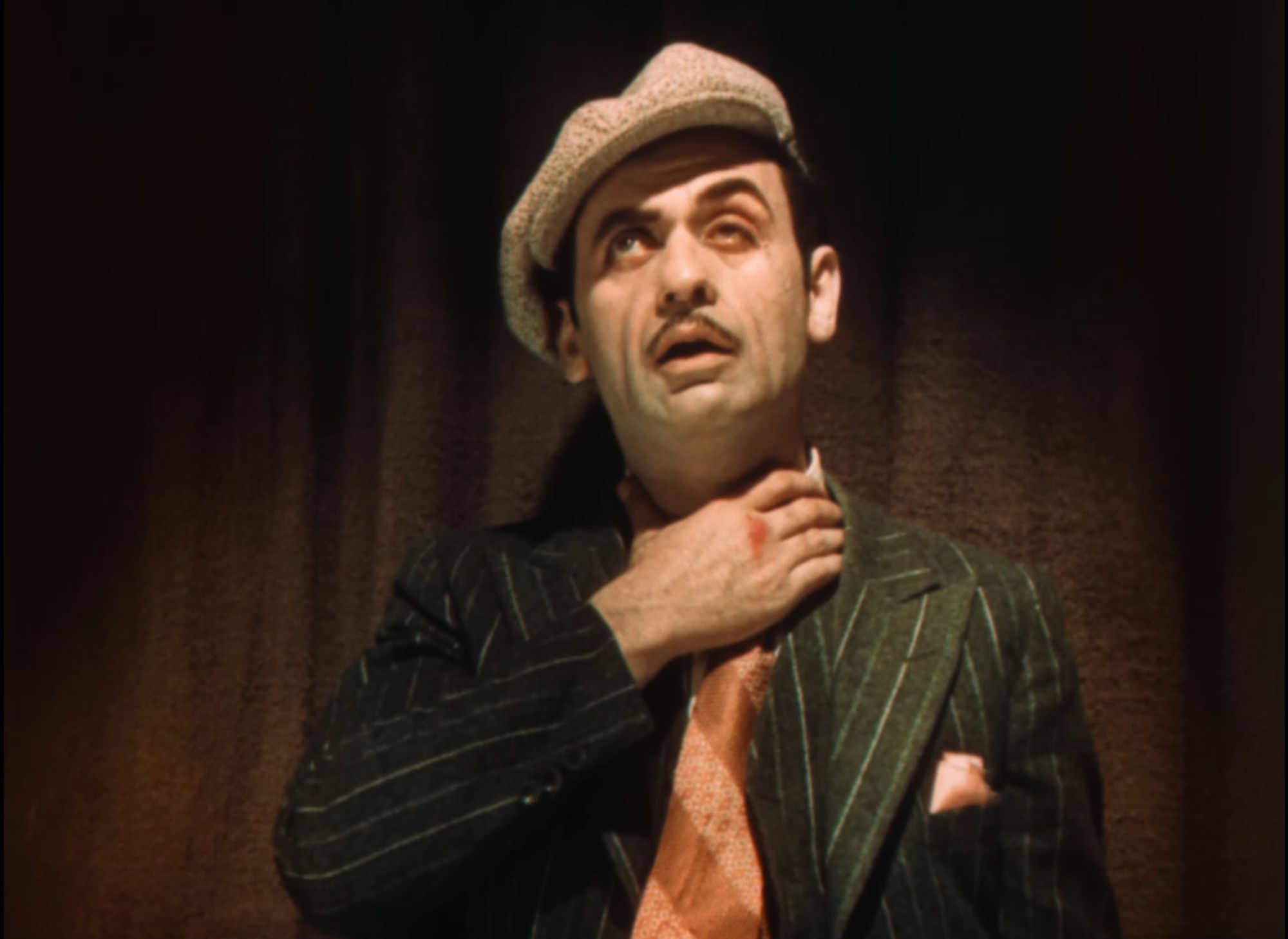 A man with a pencil moustache in a flat cap, pinstripe suit and broad striped necktie clutches his throat