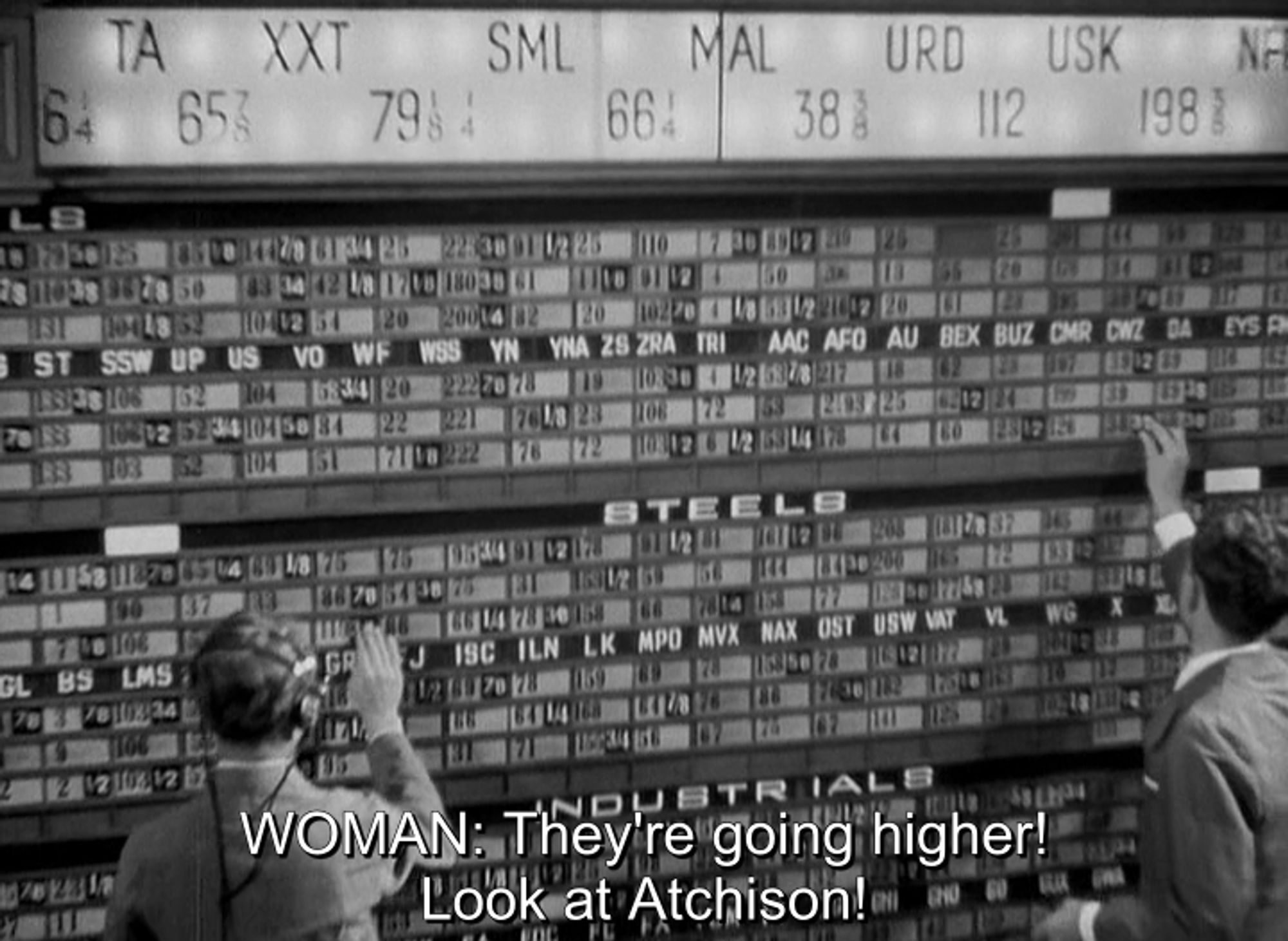 Three Cornered Moon (1933). A floor-to-ceiling display of stock price information, with a large, back-lit, scrolling ticker at the top. A couple of attendants in sport coats, one wearing a pair of headphones, are rearranging numbers on the board to reflect new info. The subtitle reads, "WOMAN: They're going higher! Look at Atchison!"