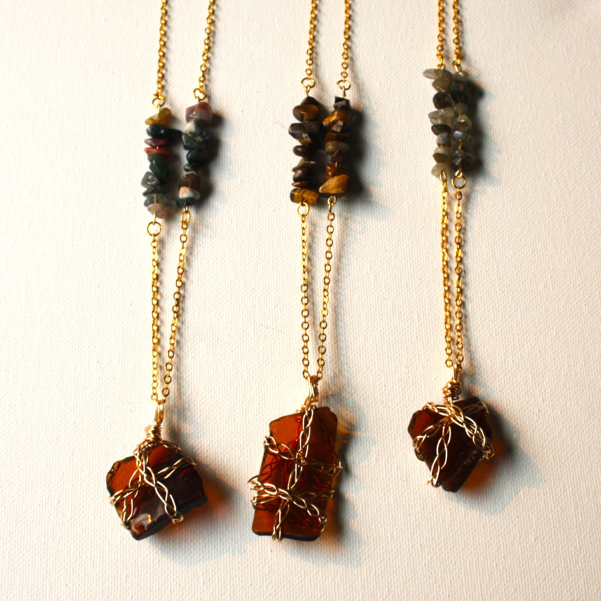 3 gold necklaces with brown sea glass wrapped in braided gold wire, their necklace chains decorated with various colored polished stone beads.