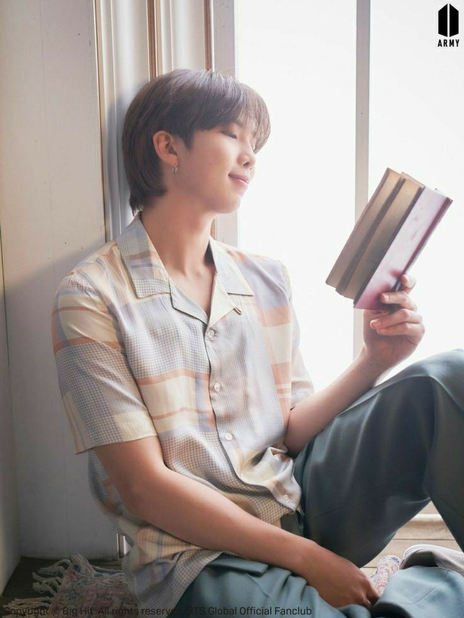 RM sits on the floor by the window reading a book. He has a smile on his face. He's wearing light blue pressed trousers, and a blue/tan/orange plaid short-sleeved button-up shirt. The floor is dark brown wood with a tapestry rug, and the walls are white.