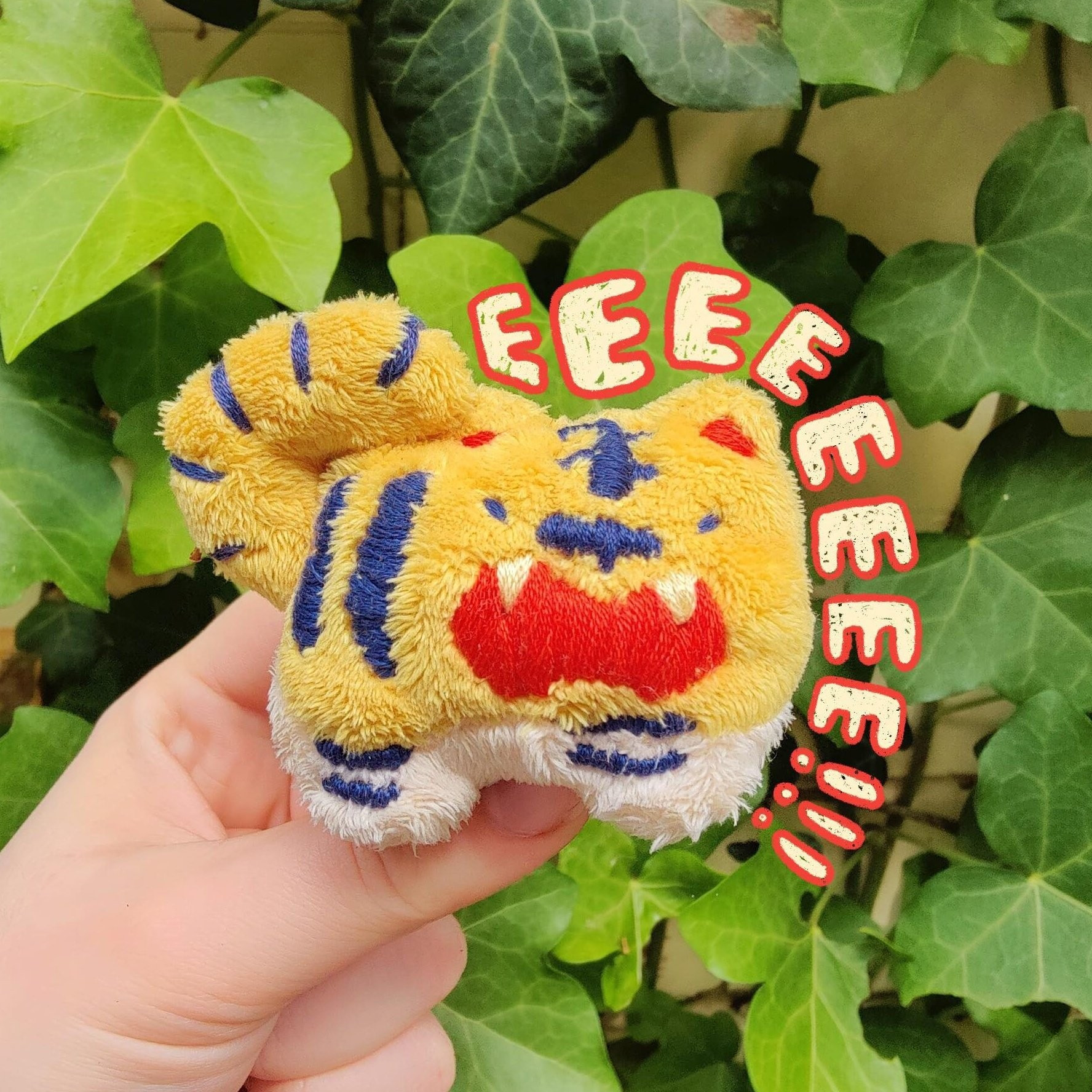 A photo of a handmade palm-sized tiger plush, very small and round and fat. He's got his mouth open in a big yell, with "EEEEEEEE!!!" being drawn around him.