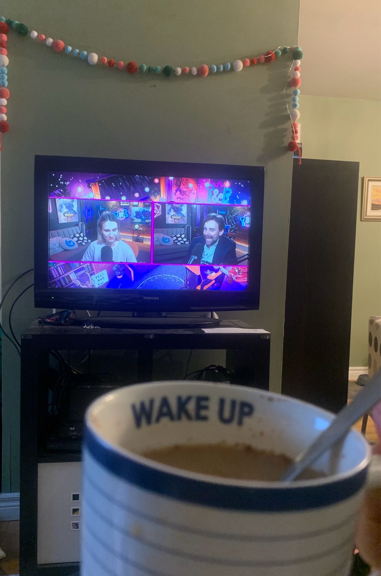 A photo of a cup of tea, with a tv screen in the background with Taliesin and Evitel’s podcast on it.