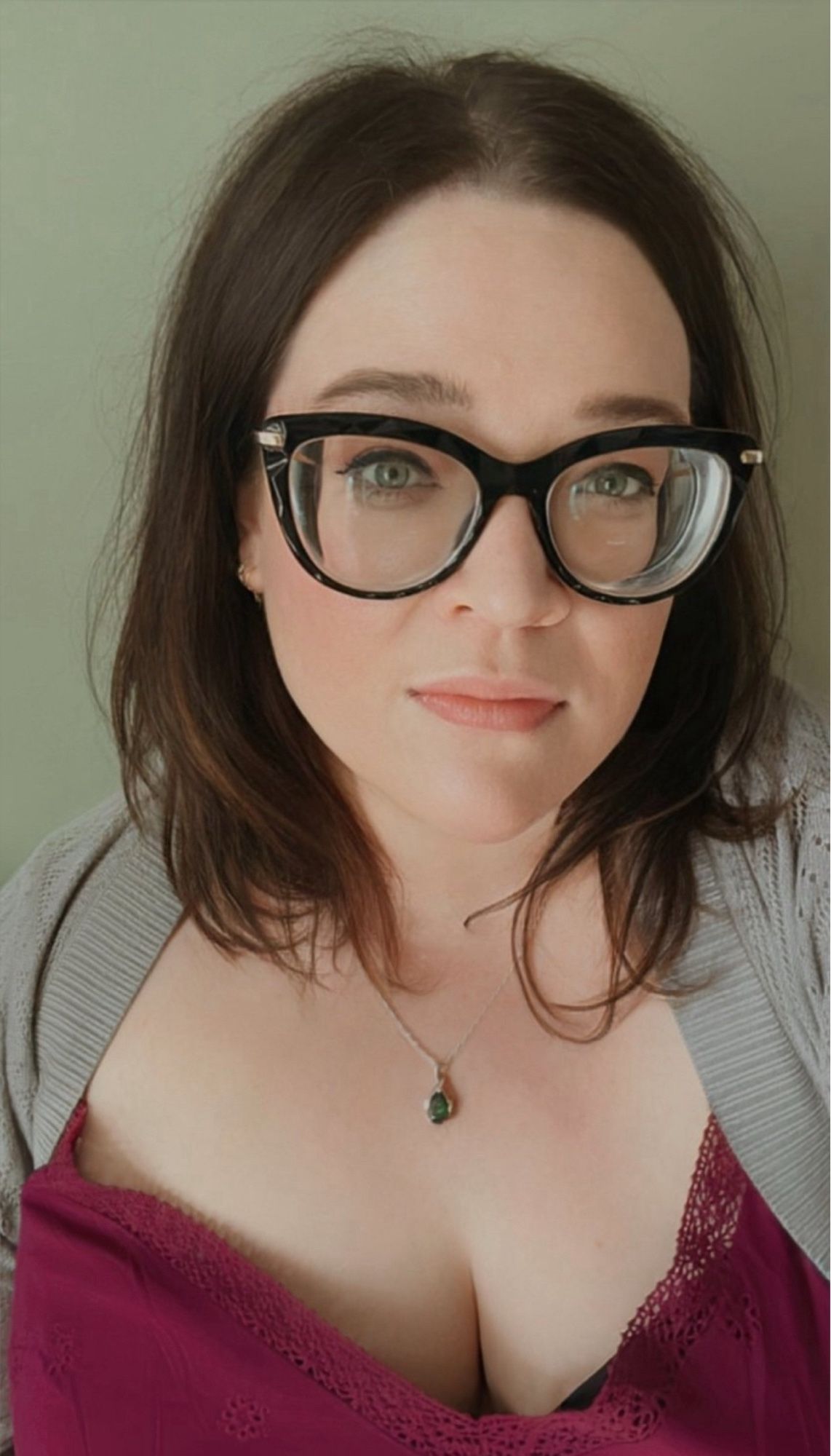 Selfie of a woman wearing large plastic black frame glasses, brown, shoulder length hair and green eyes.