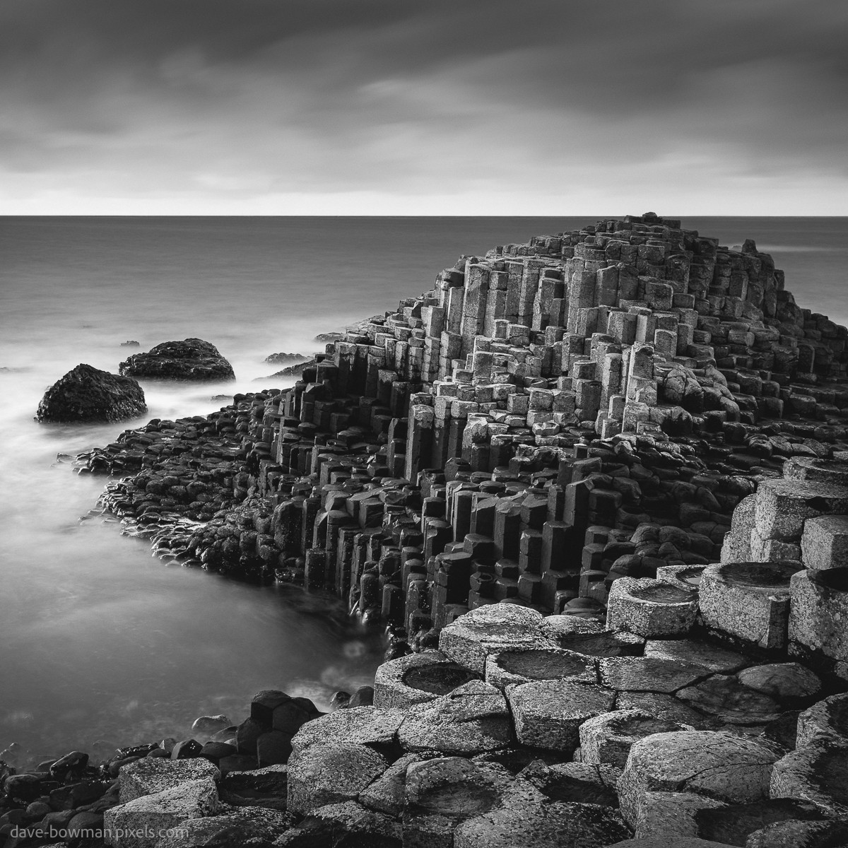 A photograph of the Giant's Causeway in County Antrim, Northern Ireland, shown in black and white. The hexagonal rock formations stretch out towards the sea, standing clearly against the sky. The waves and clouds appear smooth and blurred due to a long exposure, giving the scene a calm and quiet feeling. The image highlights the natural shape and texture of this unique coastline.