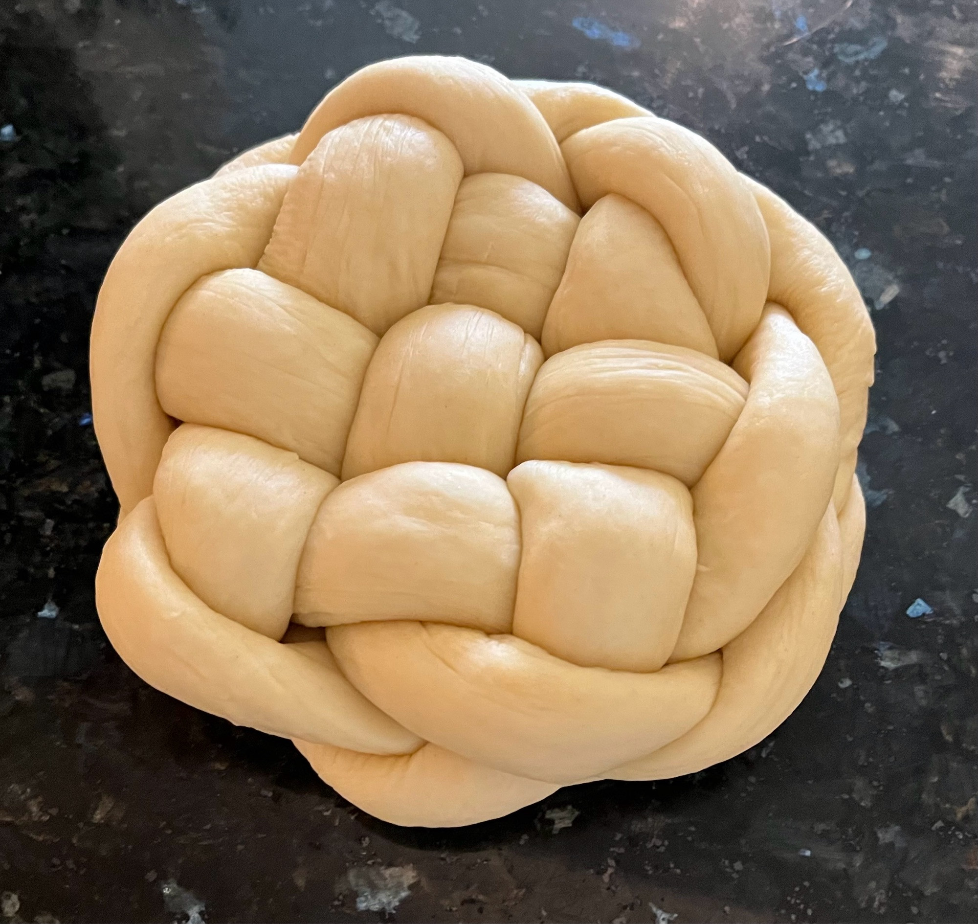 Unbaked round braided six-strand challah sitting on a black granite countertop