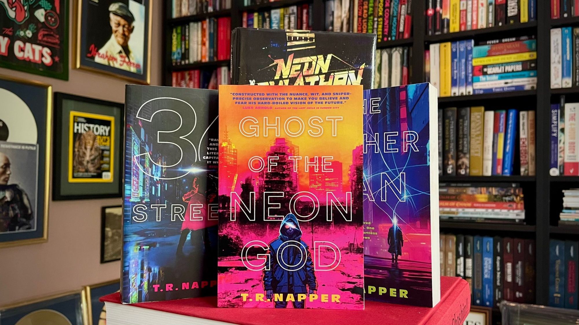 A picture showing books by TR Napper, prominently showing GHOST OF THE NEON GOD, showing a hooded figure with a cityscape behind them