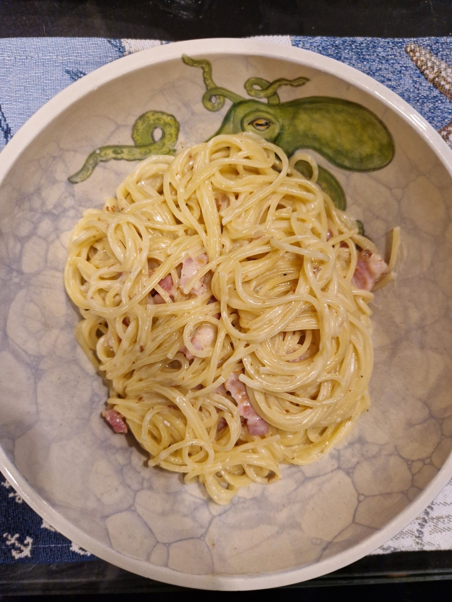 Das Bild zeigt eine Schüssel mit Spaghetti Carbonara. Die Spaghetti sind mit einer cremigen Sauce und kleinen Speckstückchen vermischt. Die Schüssel hat ein dekoratives Design mit einem grünen Oktopus auf der linken Seite. Der Hintergrund der Schüssel hat ein marmoriertes Muster. Die Schüssel steht auf einem blauen Tischset mit einem maritimen Muster.