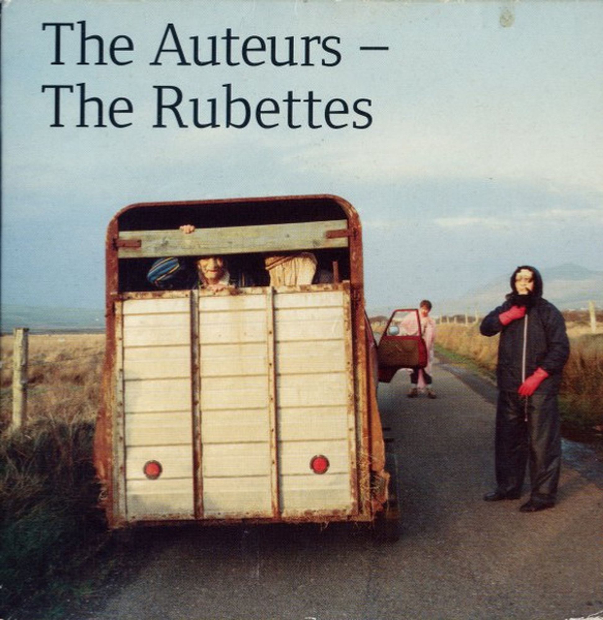 The sleeve of The Rubettes by The Auteurs. A photo of the back of a horse box parked on a country road. An old woman, possibly masked, looks out of the back. A man in a black parka waterproof trousers and red gloves, possibly also masked, stands by the vehicle. The scene feels oddly disturbing.