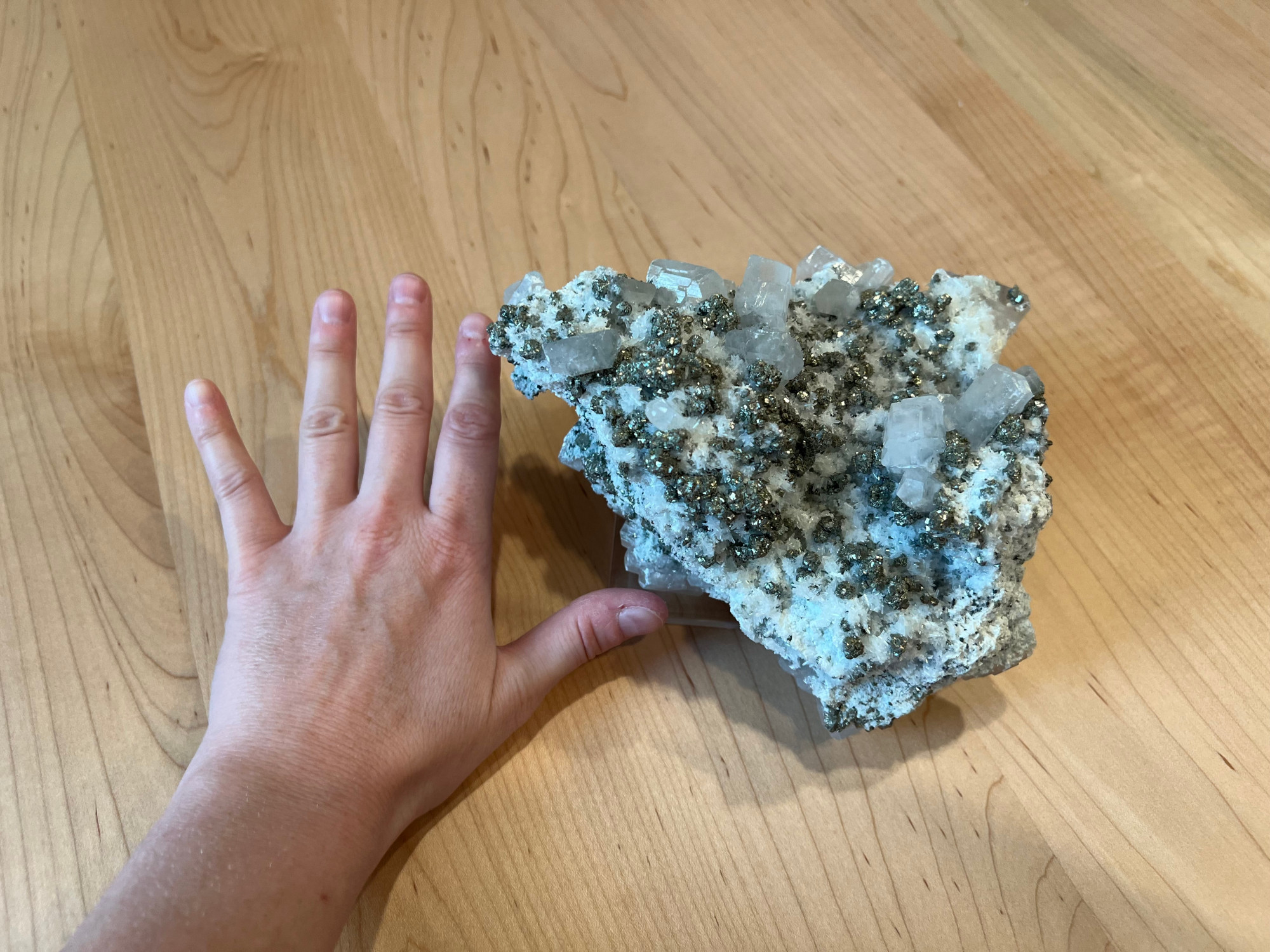 A huge mineral specimen dotted with chalcopyrite, pyrite, and sphalerite in addition to white calcite and quartz crystals. There is a hand to the left for scale (the rock is about the size of the hand with fingers spread if not larger).