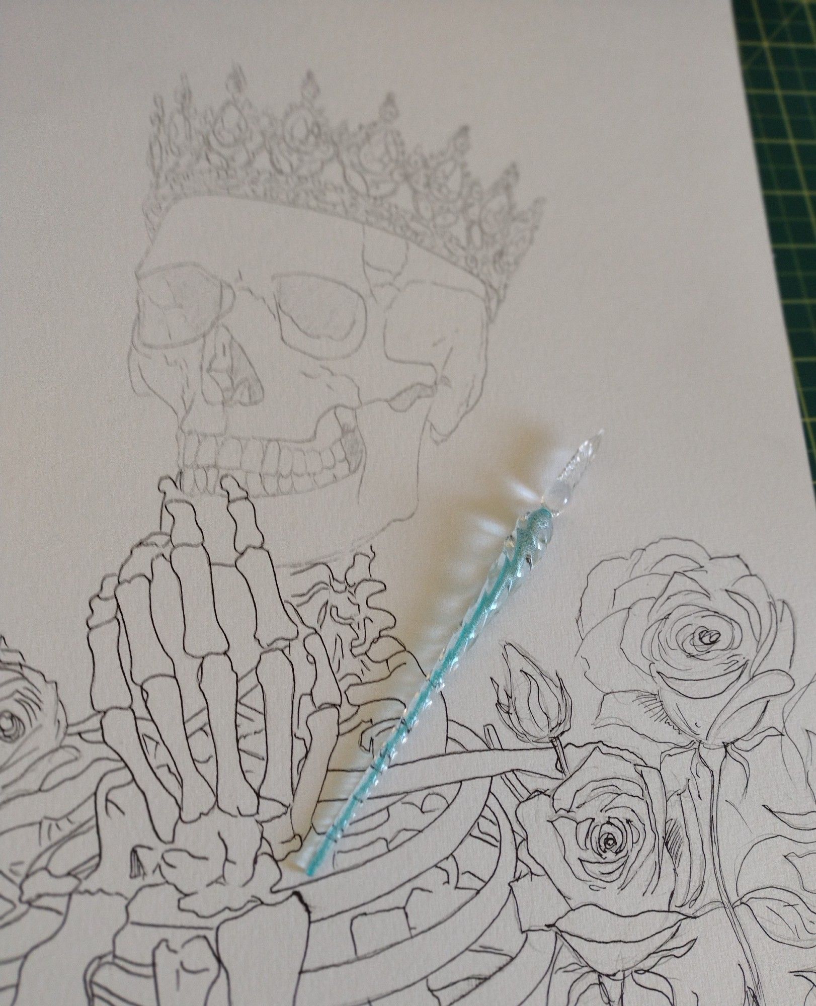 A turquoise glass dip pen on top of an incompete drawing of a skeleton and roses.